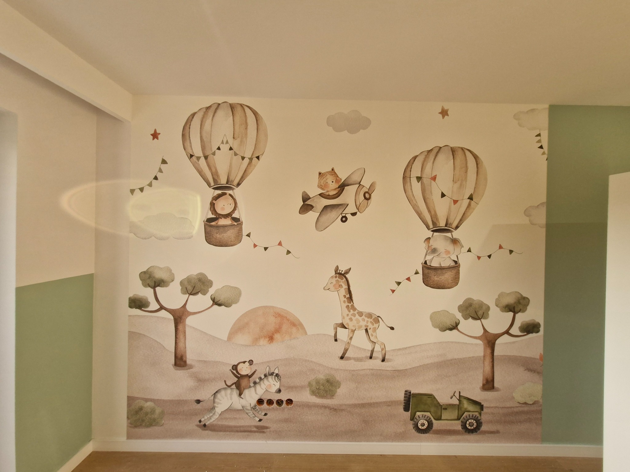 Pokój dziecięcy z tapetą w stylu safari: żyrafa, słoń w balonie, małpka na zebrze i jeep na tle drzew i słońca. Stonowana kolorystyka, przytulna atmosfera.