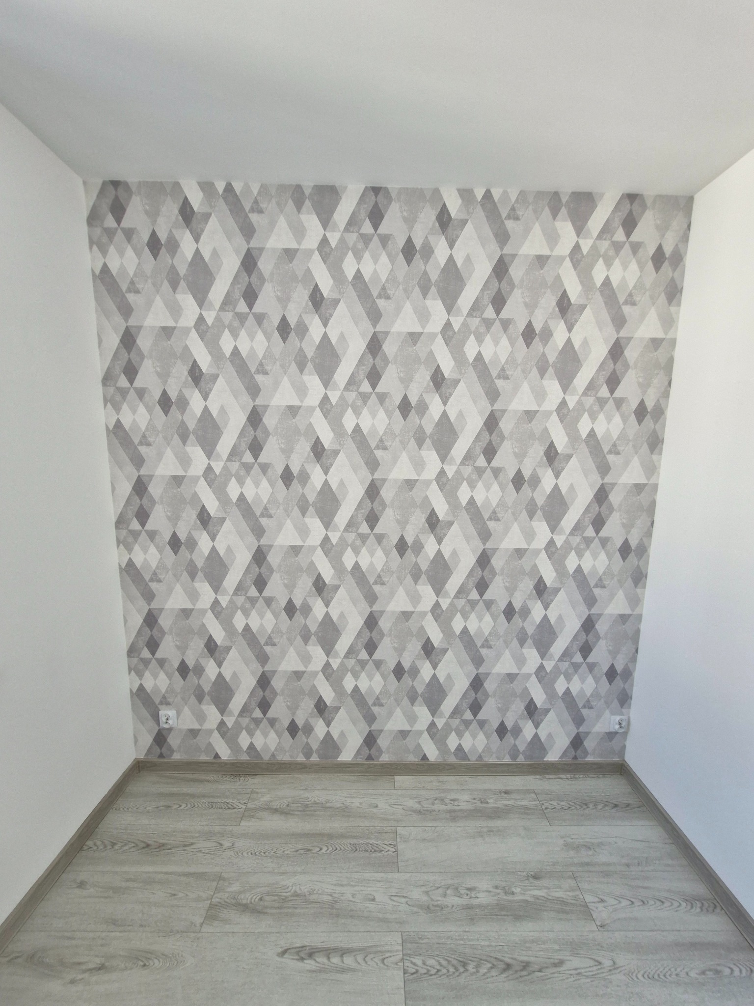 Pokój po remoncie: ściana z szarą tapetą w geometryczne wzory, jasna podłoga, białe ściany boczne. Widoczne gniazdka elektryczne.