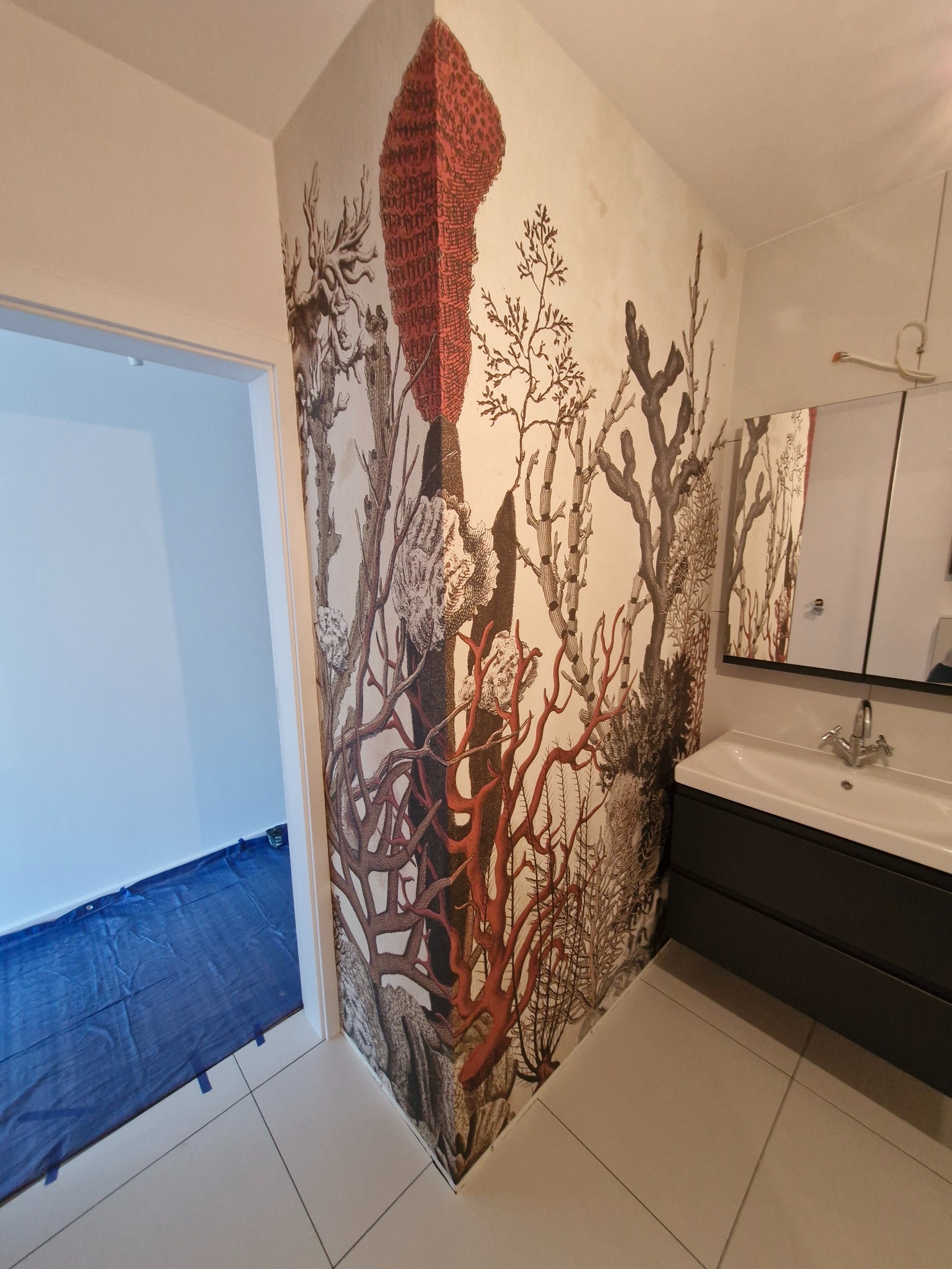 Tapeta w łazience z motywem koralowców i roślin, na podłodze folia ochronna. Widoczna umywalka i lustro. Nietypowy wzór tapety.