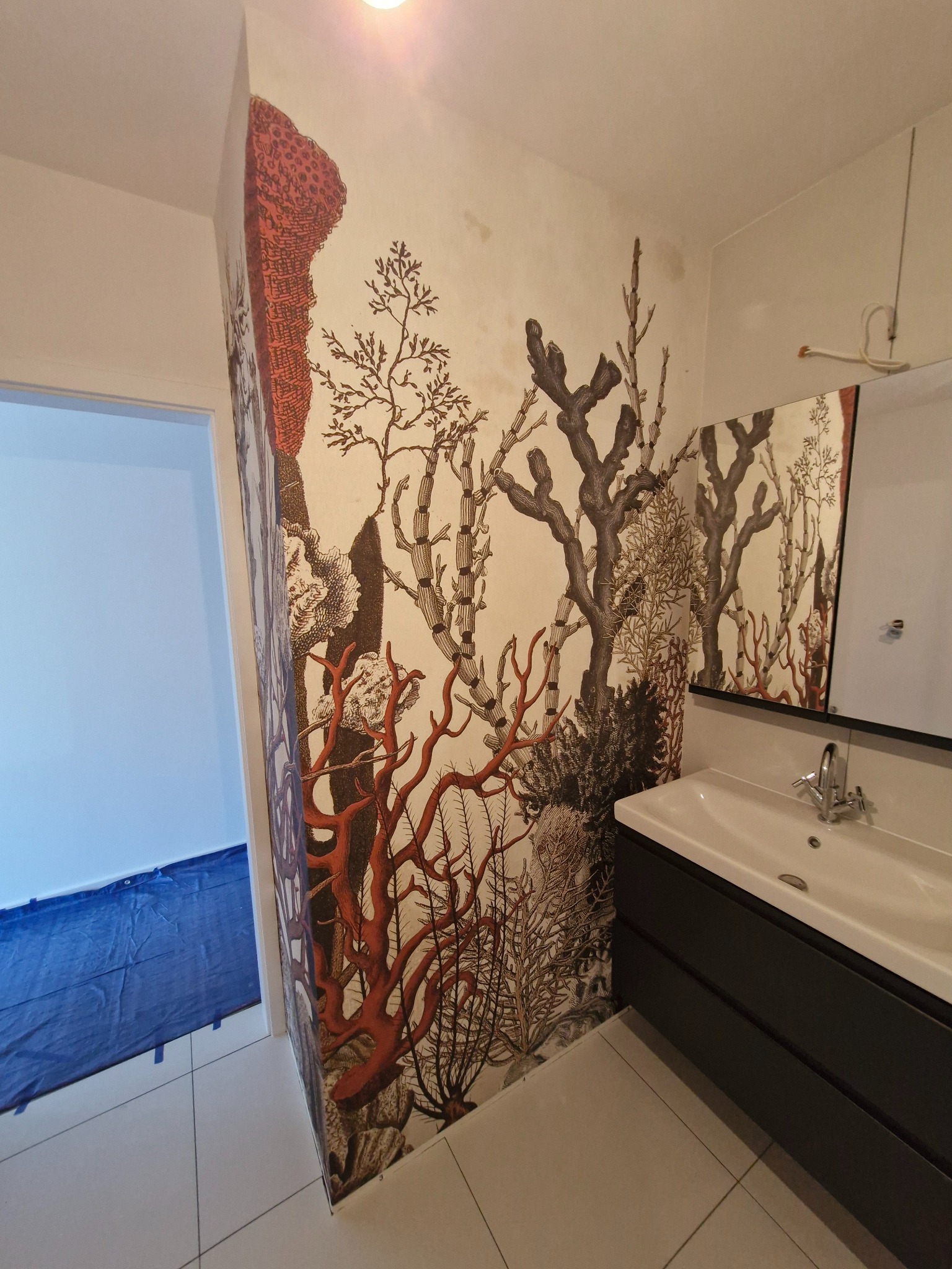 Wzorzysta tapeta w łazience z umywalką. Motyw roślinny w odcieniach brązu i czerwieni tworzy oryginalny wystrój. Widoczny fragment zabezpieczonej podłogi.