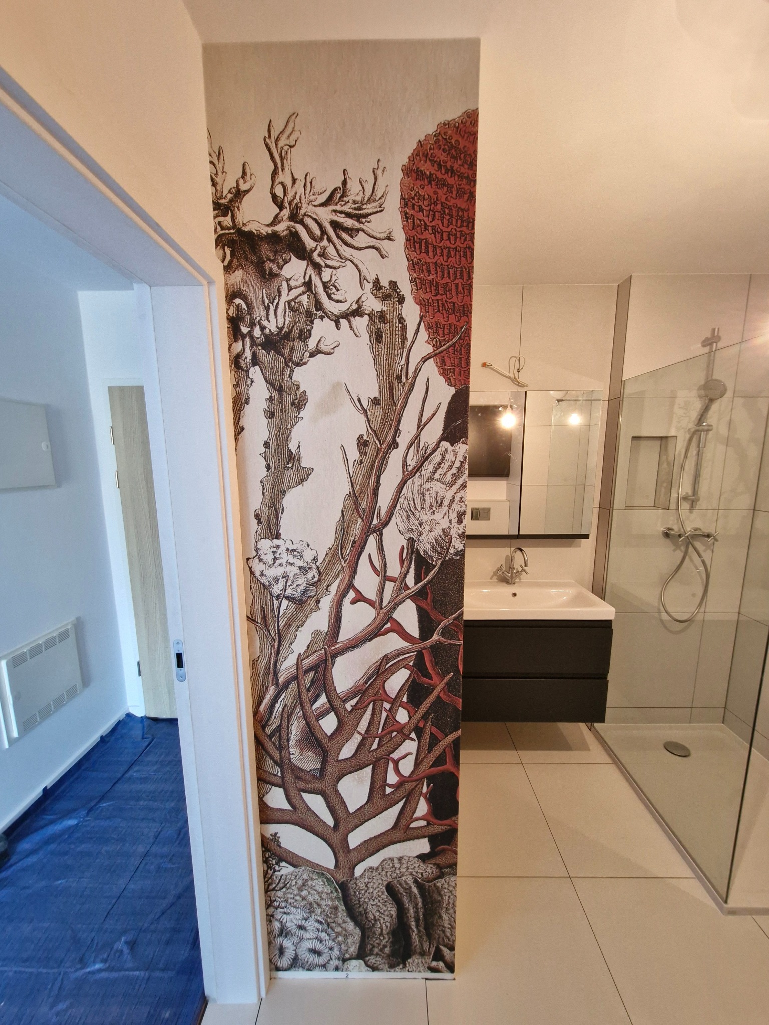 Wykończona ściana w łazience z unikalną tapetą w motywy roślinne, widok na umywalkę i prysznic. Na podłodze jasne płytki.