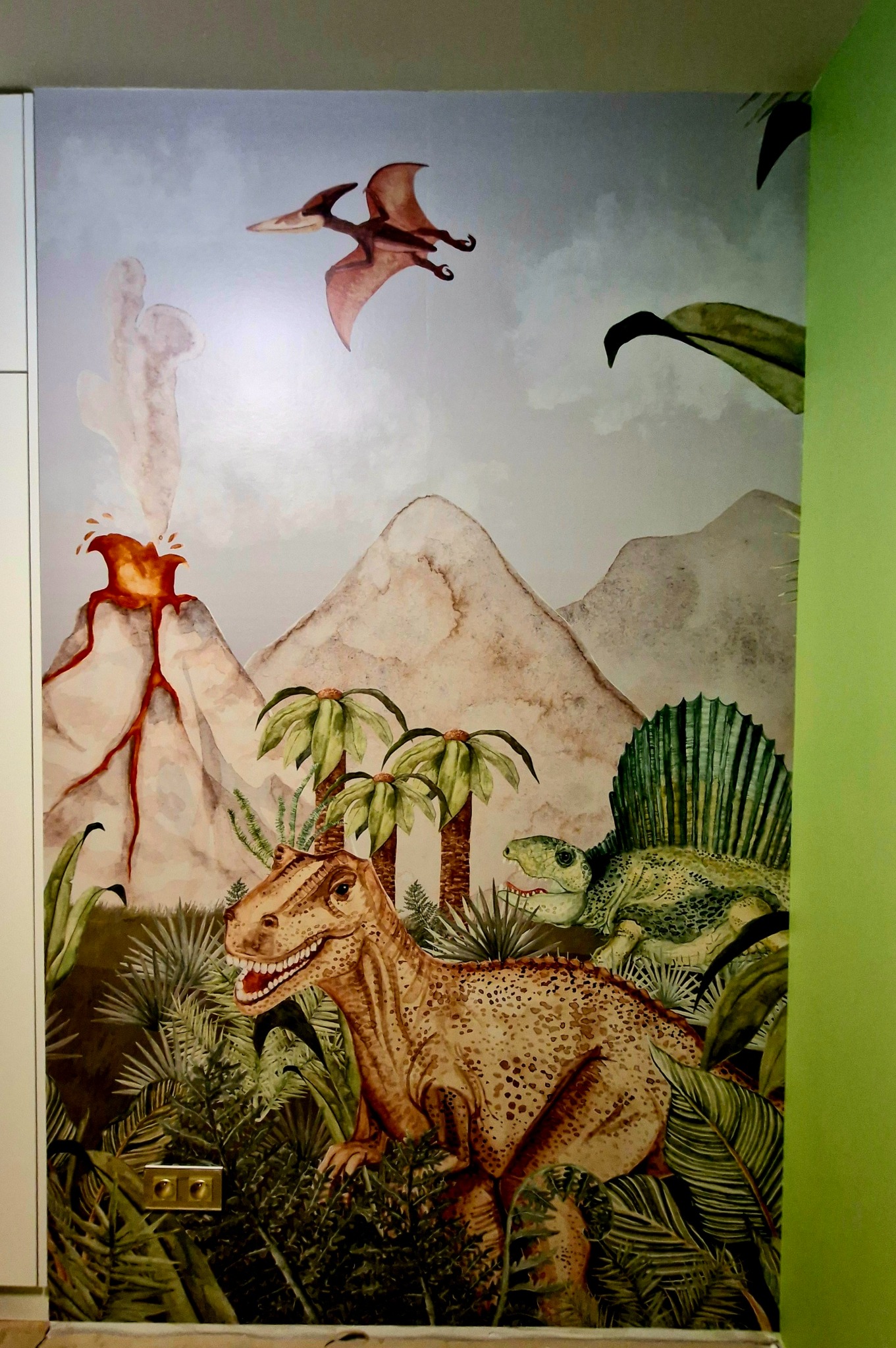 Dinozaury i wulkan na tapecie w pokoju dziecięcym. Dekoracja ściany z motywem prehistorycznym, widoczne gniazdka elektryczne na tle dżungli.