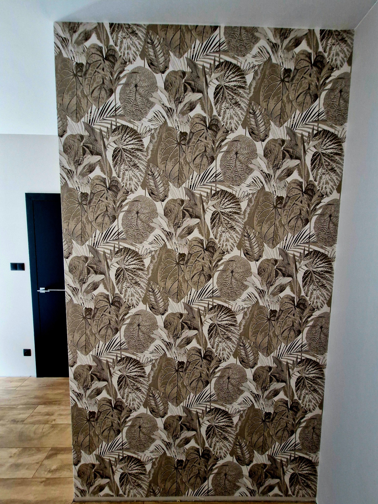 Ściana z tapetą w motywy liściaste w odcieniach brązu i bieli, widoczna część podłogi i fragment czarnych drzwi w tle, po remoncie.