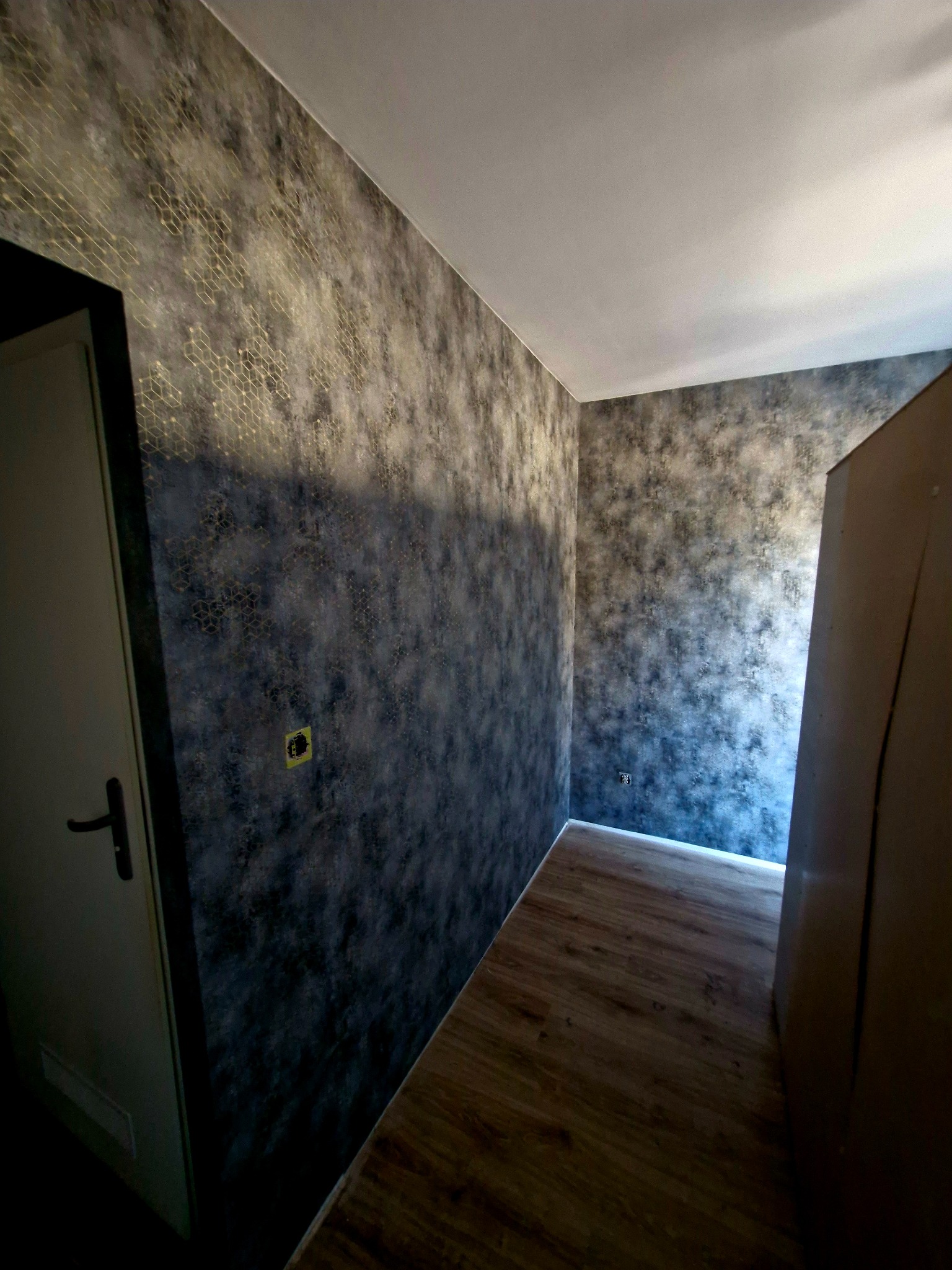 Pokój po odświeżeniu: ściany zdobi tapeta w odcieniach szarości z delikatnym, geometrycznym wzorem w kolorze złota. Widoczna część drzwi i fragment podłogi.