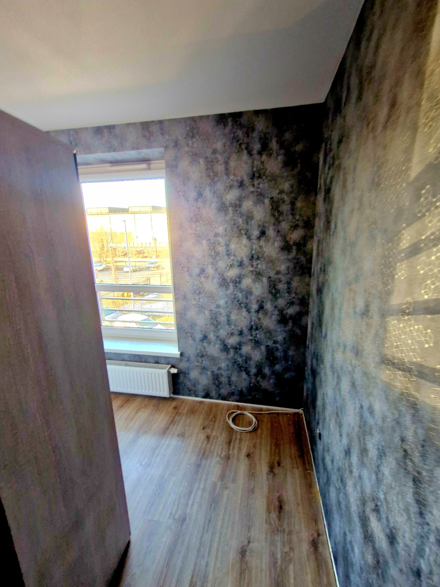 Pokój po tapetowaniu w ciemnych odcieniach szarości i brązu. Widoczne panele podłogowe, okno z widokiem na zewnątrz i fragment niedomkniętych drzwi.