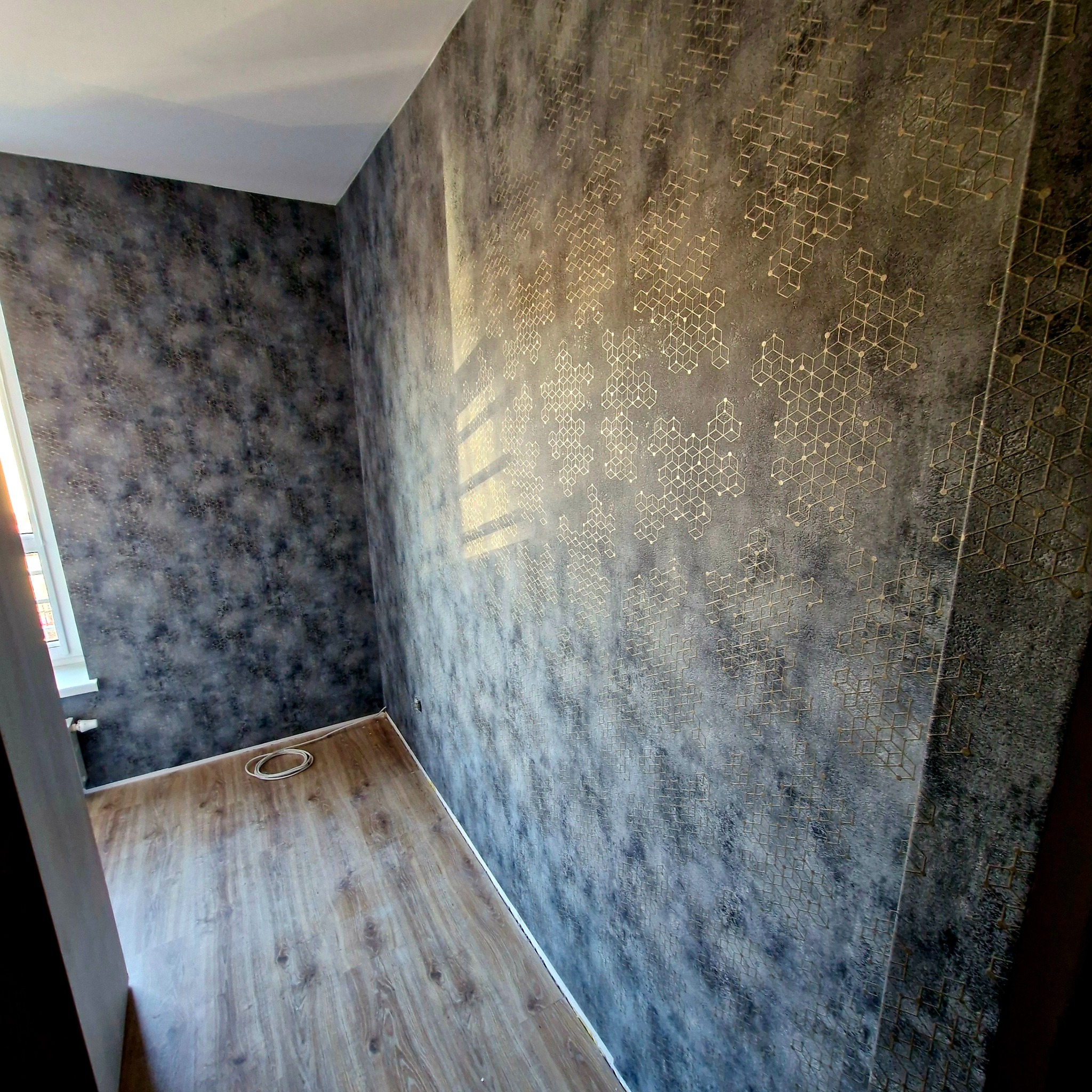 Pokój po dekoracji ścian: szara tapeta z geometrycznym, złotym wzorem. Podłoga wyłożona panelami imitującymi drewno. Widok na narożnik pokoju.