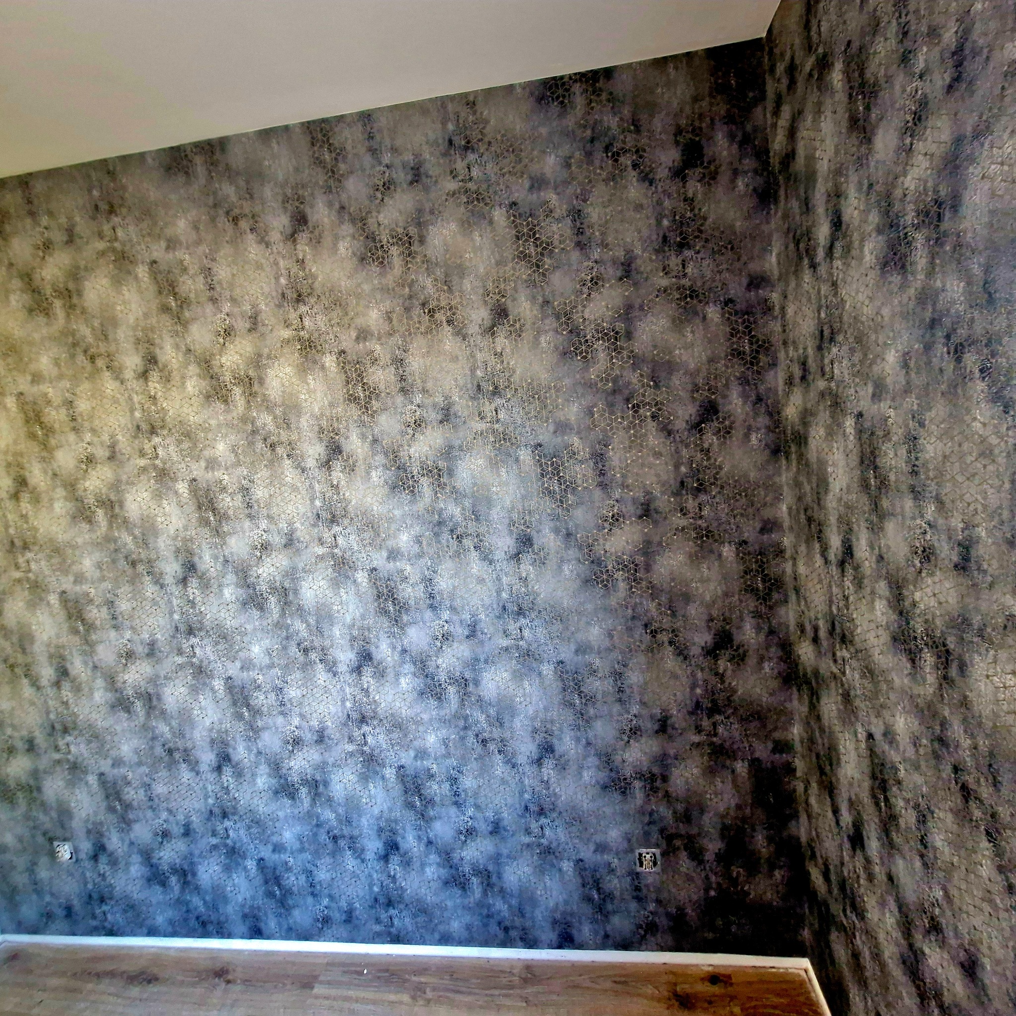 Ściana z tapetą w odcieniach szarości i srebra z geometrycznym wzorem. Widoczne gniazdka elektryczne i fragment podłogi z drewnianymi panelami.