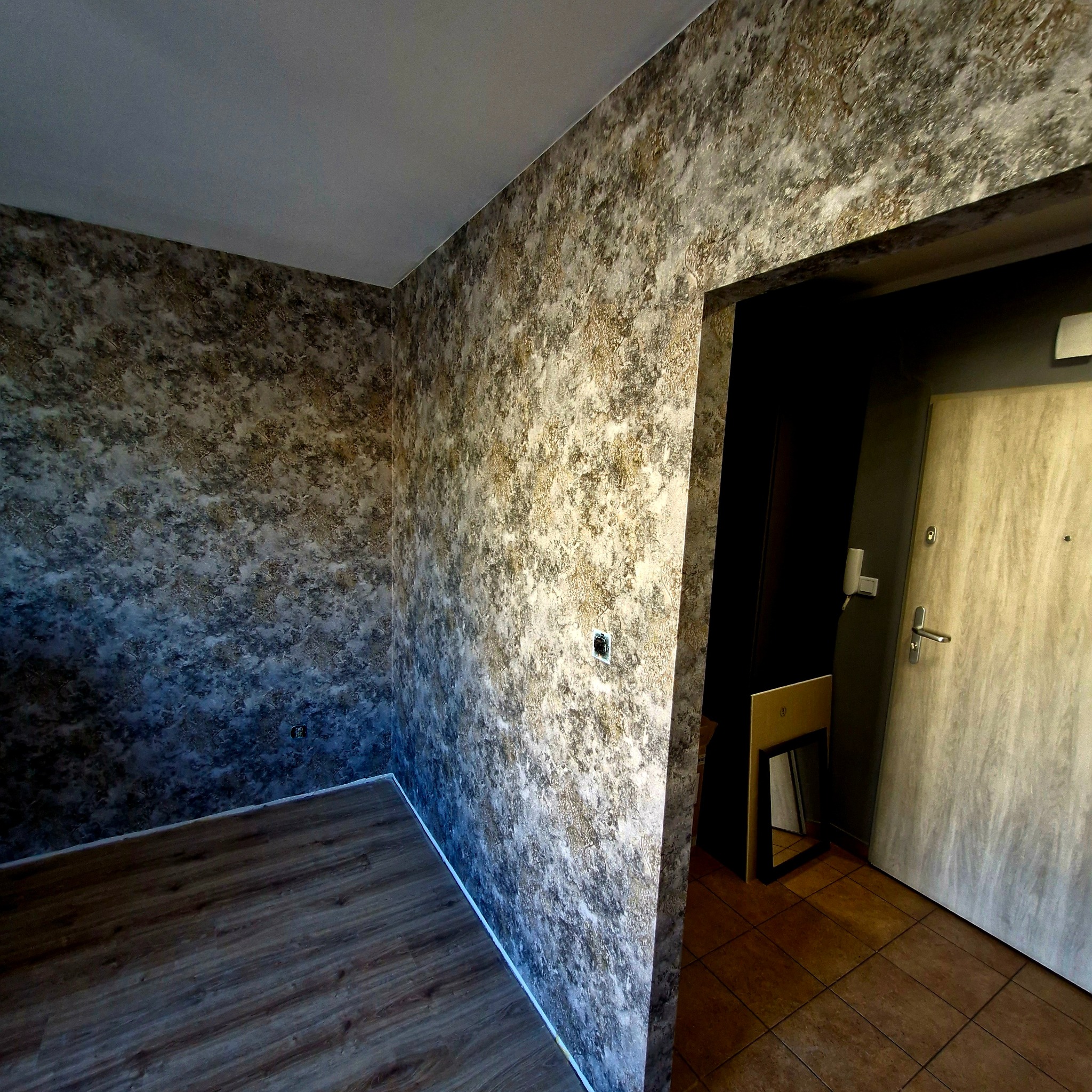 Pokój po malowaniu ścian z efektem dekoracyjnym, widoczne wykończone ściany w odcieniach brązu i beżu, podłoga w panelach. Widoczne drzwi wejściowe do mieszkania.