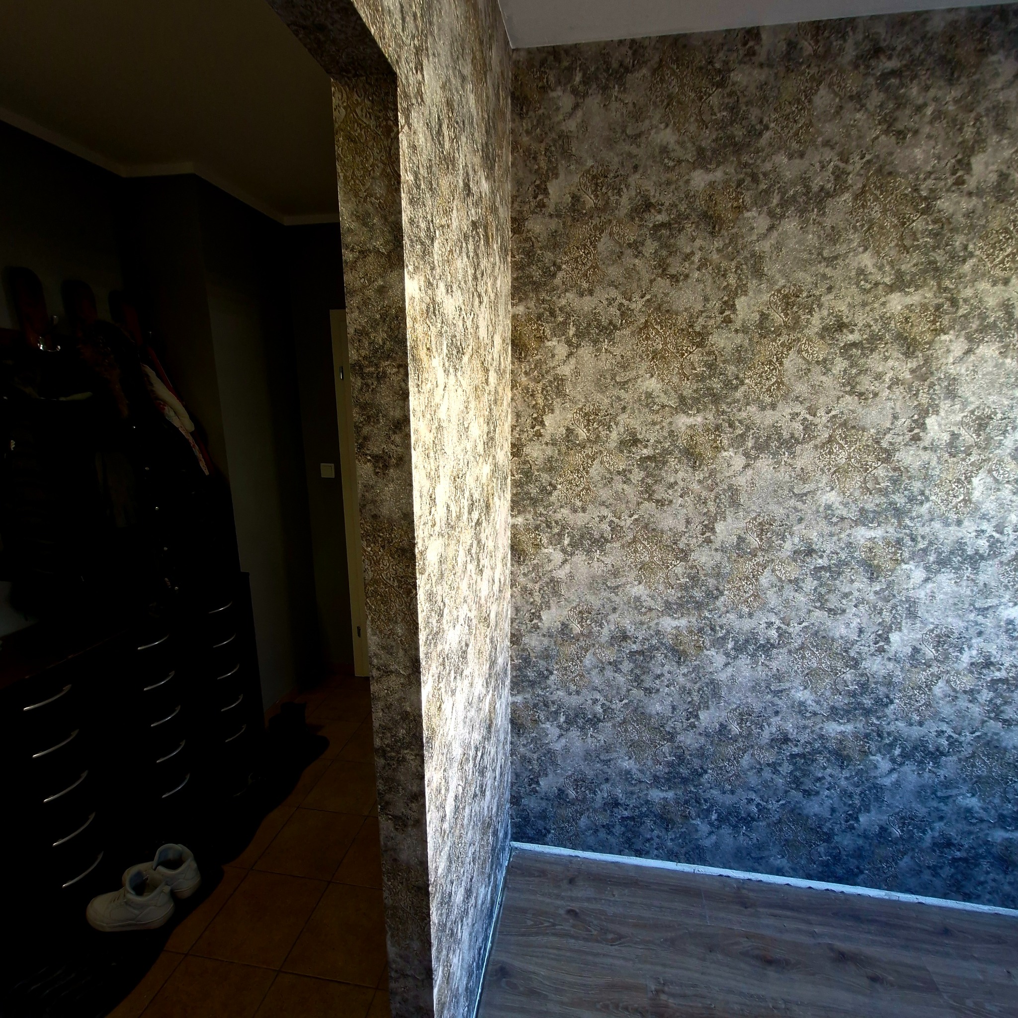 Dekoracyjna ściana z efektem tapety w odcieniach szarości i złota, widoczna z perspektywy narożnika. W tle ciemne wnętrze z widoczną szafą i obuwiem.