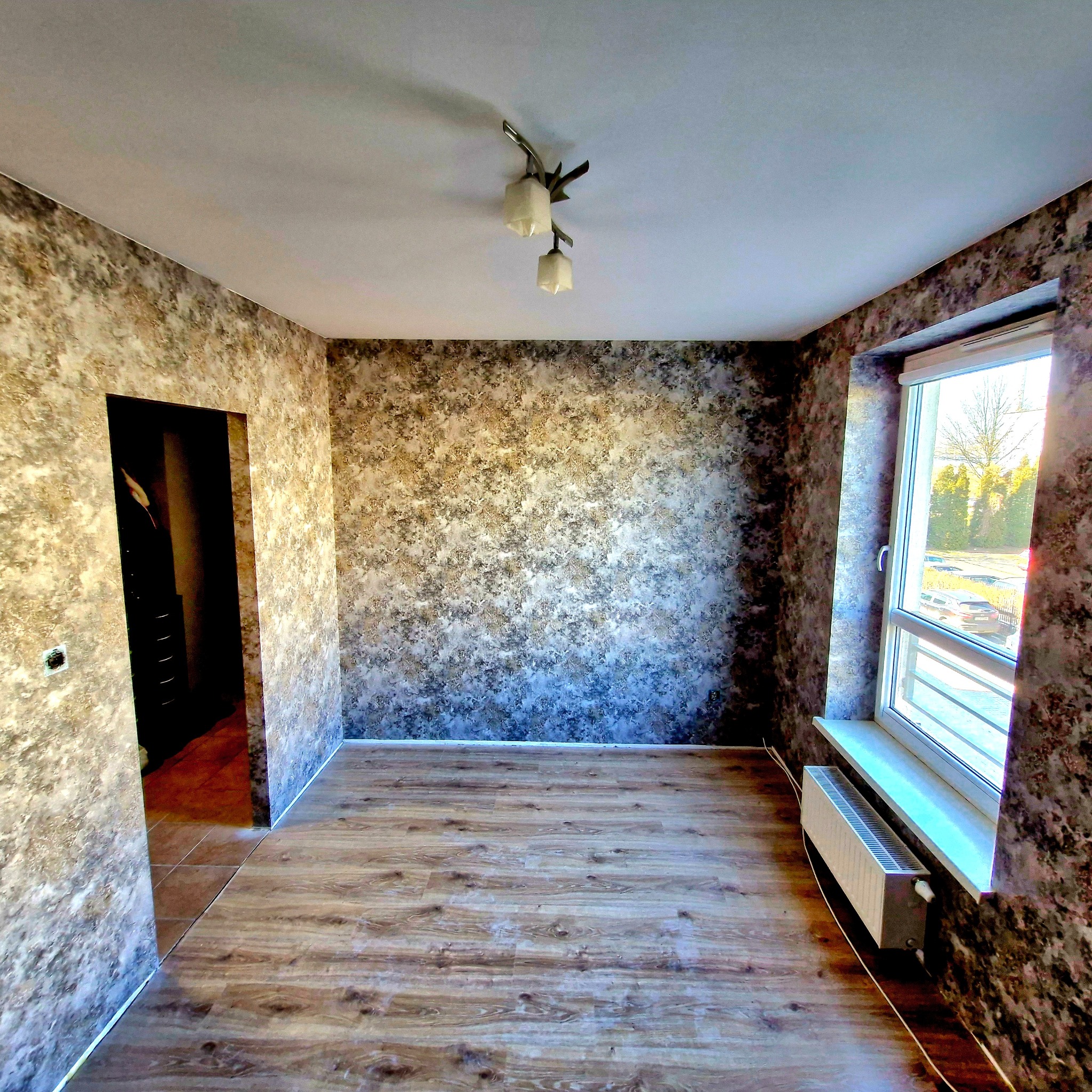 Wykończone wnętrze: ściany pokryte tapetą w odcieniach brązu i beżu, drewniana podłoga, okno z widokiem na zieleń. Lampa sufitowa i grzejnik pod oknem.