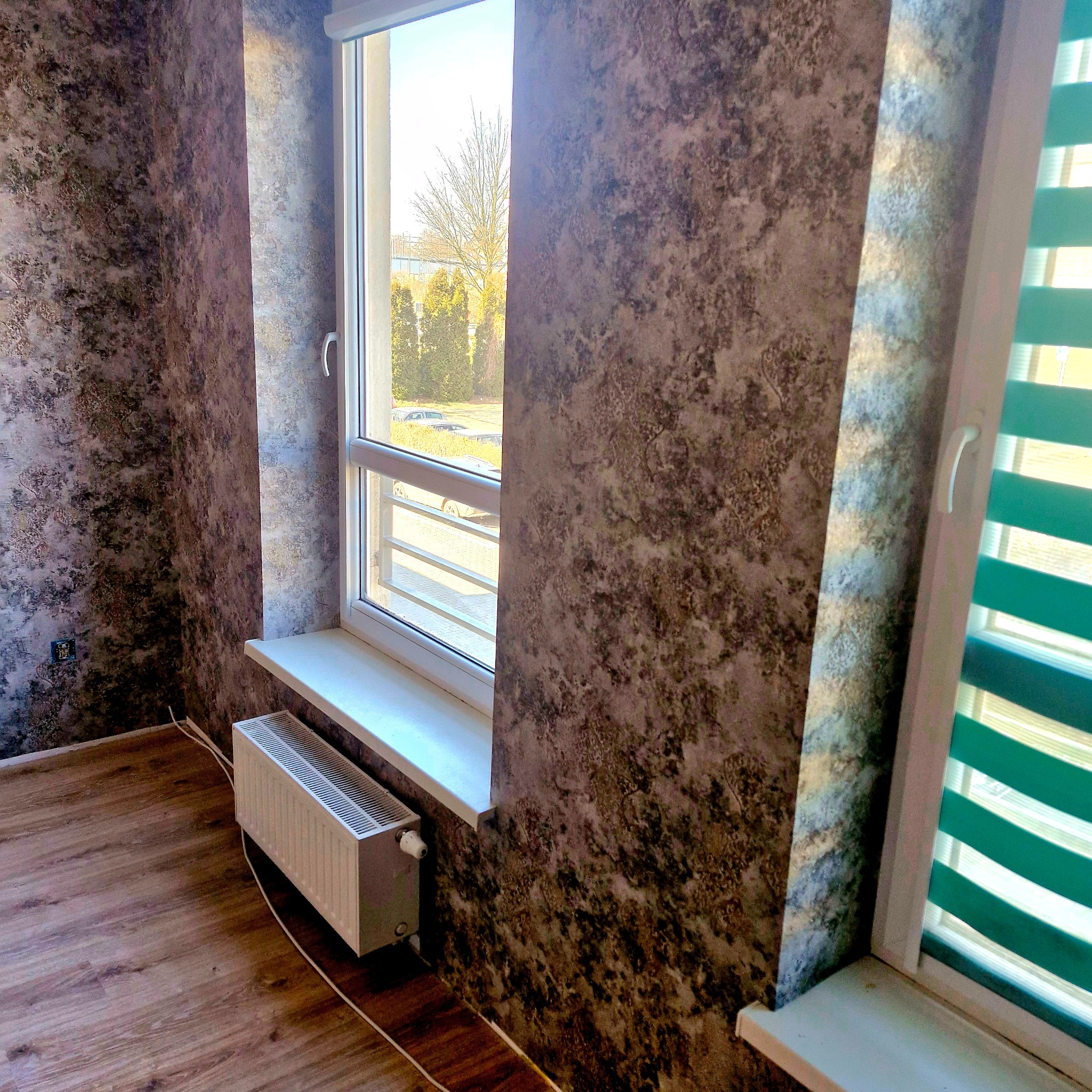 Pokój po tapetowaniu z brązowo-szarą tapetą o wzorze przypominającym kamień. Widoczne okno z roletami w kolorze turkusowym i grzejnik pod oknem. Drewniana podłoga.
