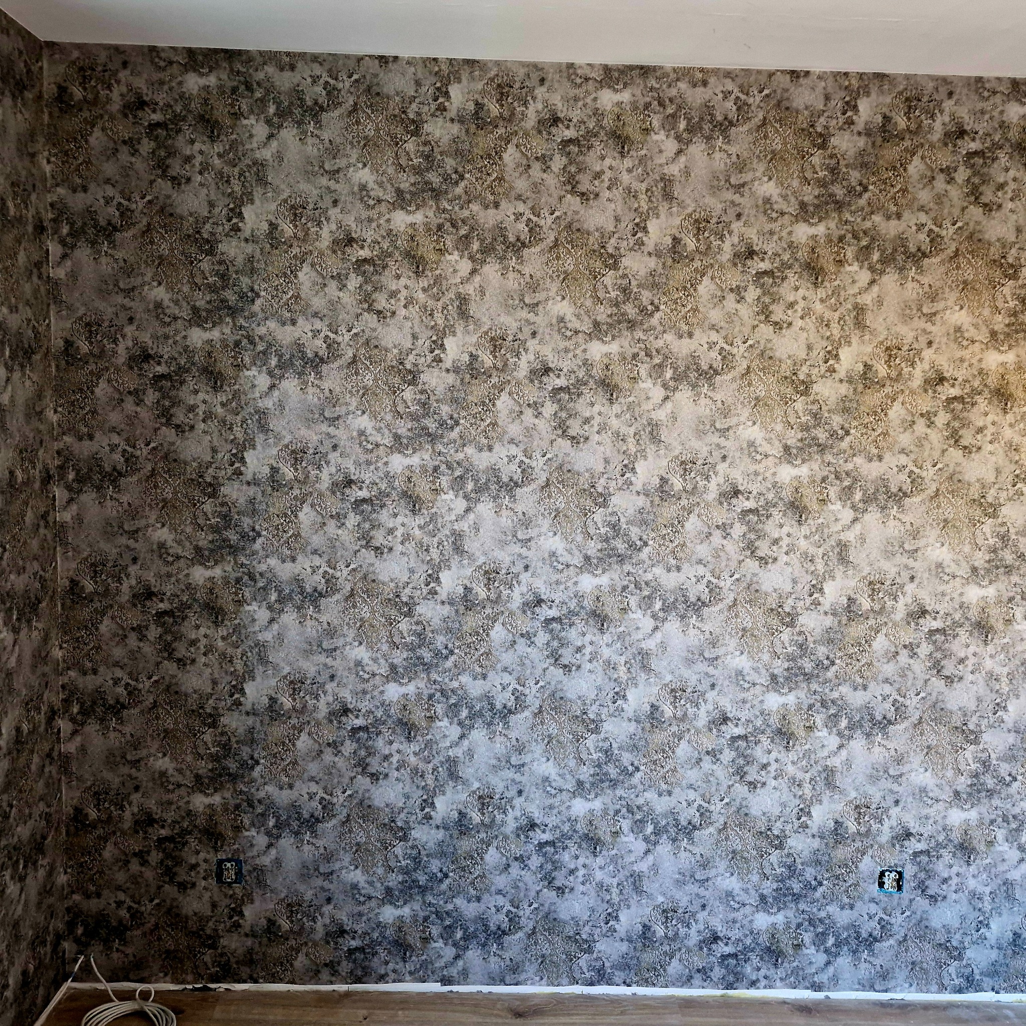 Ściana po nałożeniu dekoracyjnej tapety w odcieniach szarości i złota z subtelnym, ornamentowym wzorem. Widoczne gniazdka elektryczne i fragment podłogi.