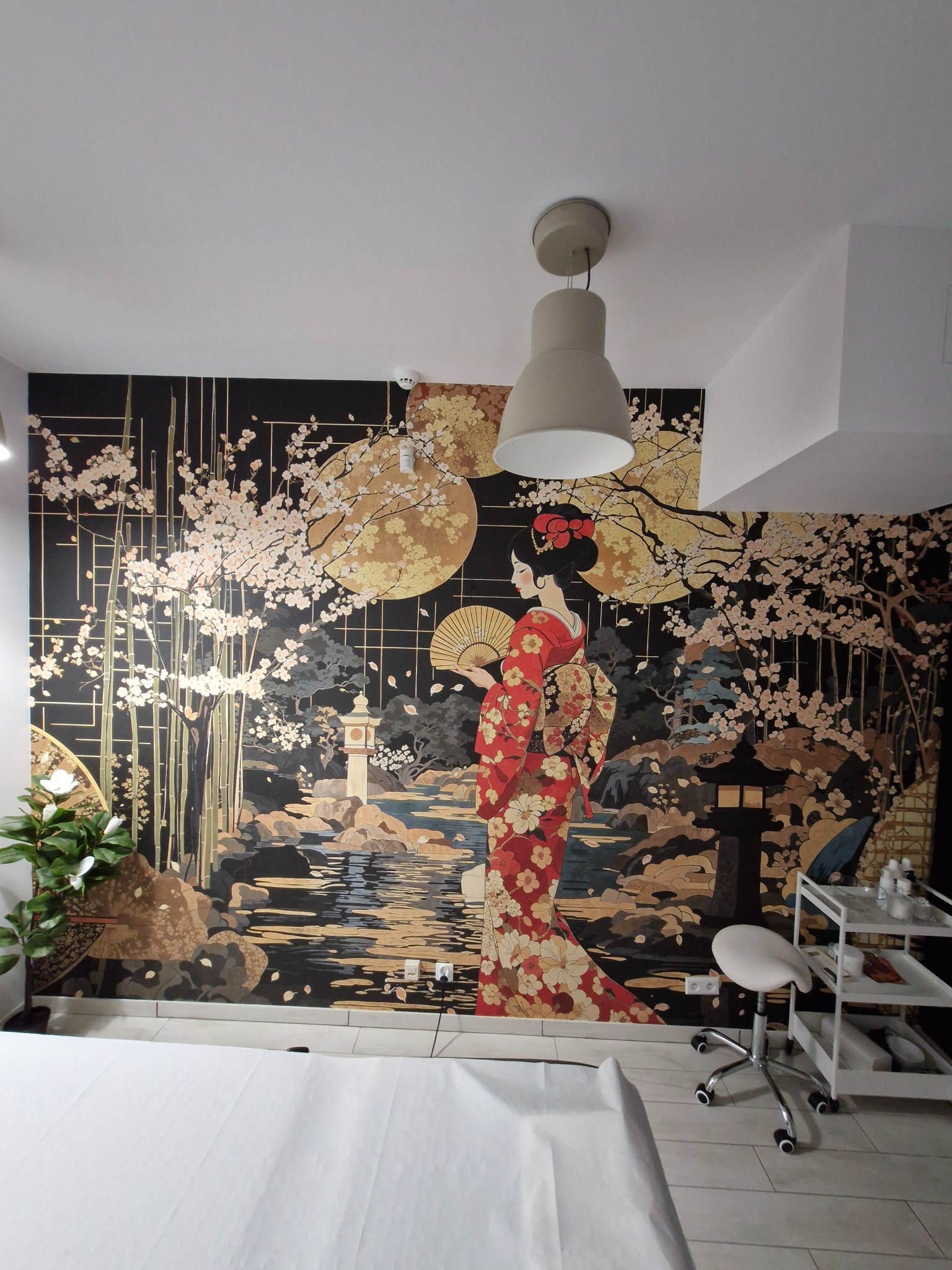 Ściana z fototapetą w stylu japońskim, przedstawiająca gejszę, bambusy i kwitnące wiśnie. Wnętrze salonu kosmetycznego z białym stołem i krzesłem.