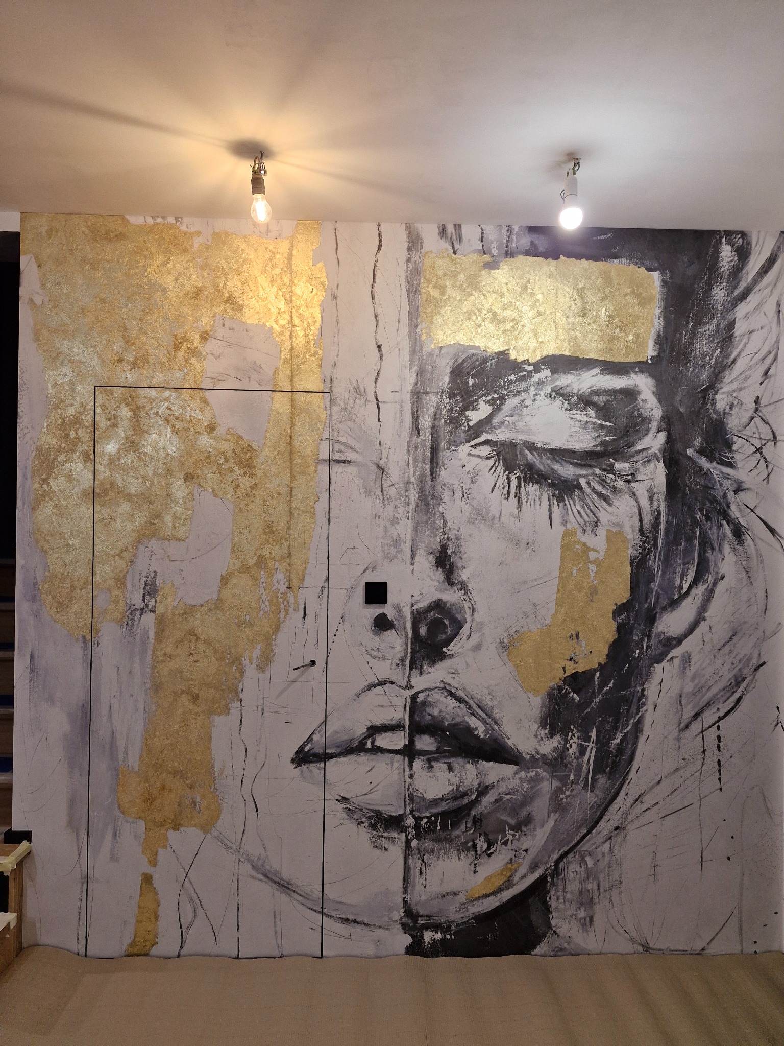 Abstrakcyjna dekoracja ściany z motywem twarzy i złotymi akcentami, oświetlona dwoma punktowymi lampami. Unikalny design wnętrza.