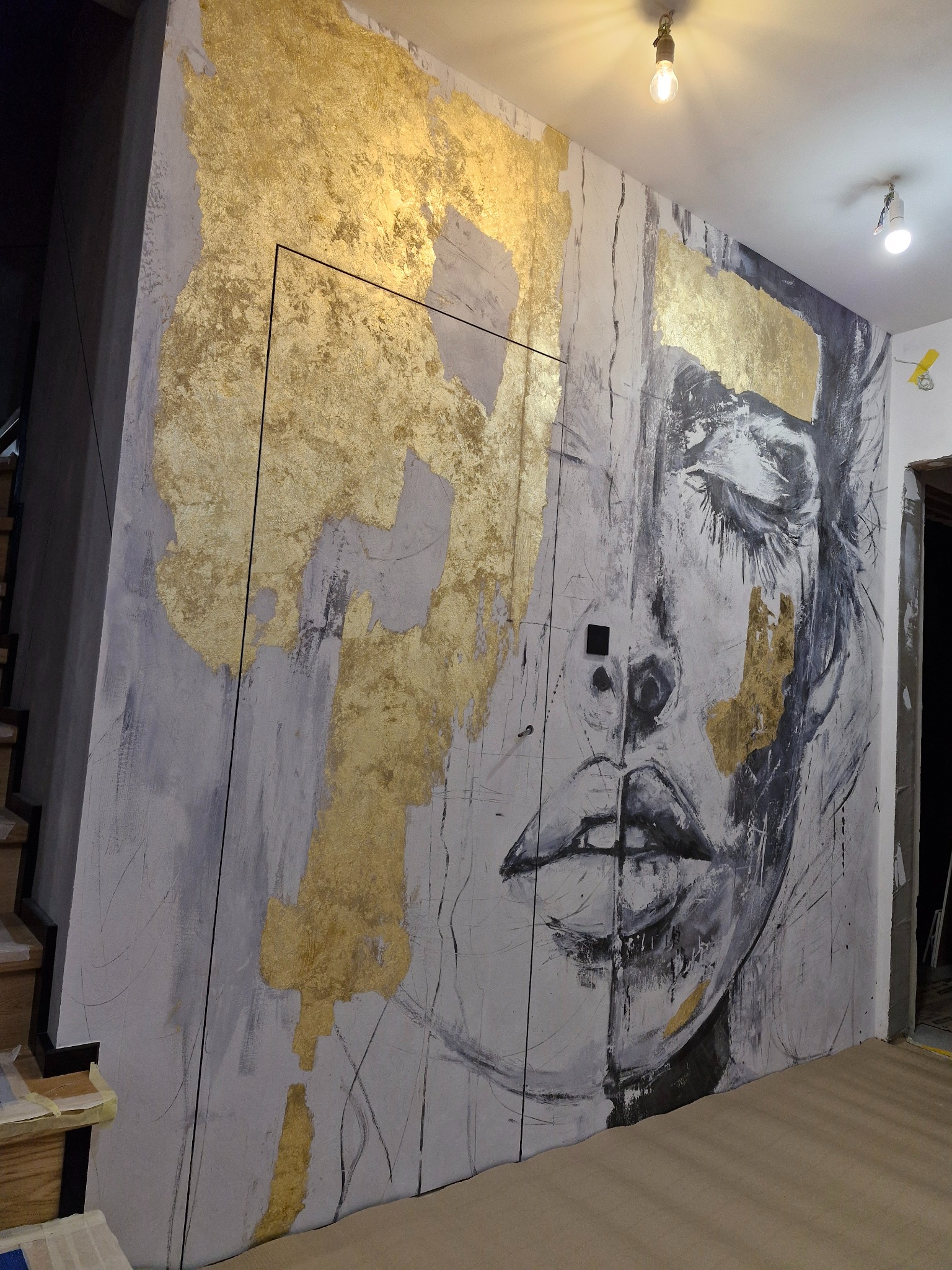 Artystyczne malowanie ściany z motywem twarzy i złotymi akcentami. Widoczne niedoskonałości dodają charakteru. Oświetlenie podkreśla fakturę farby.