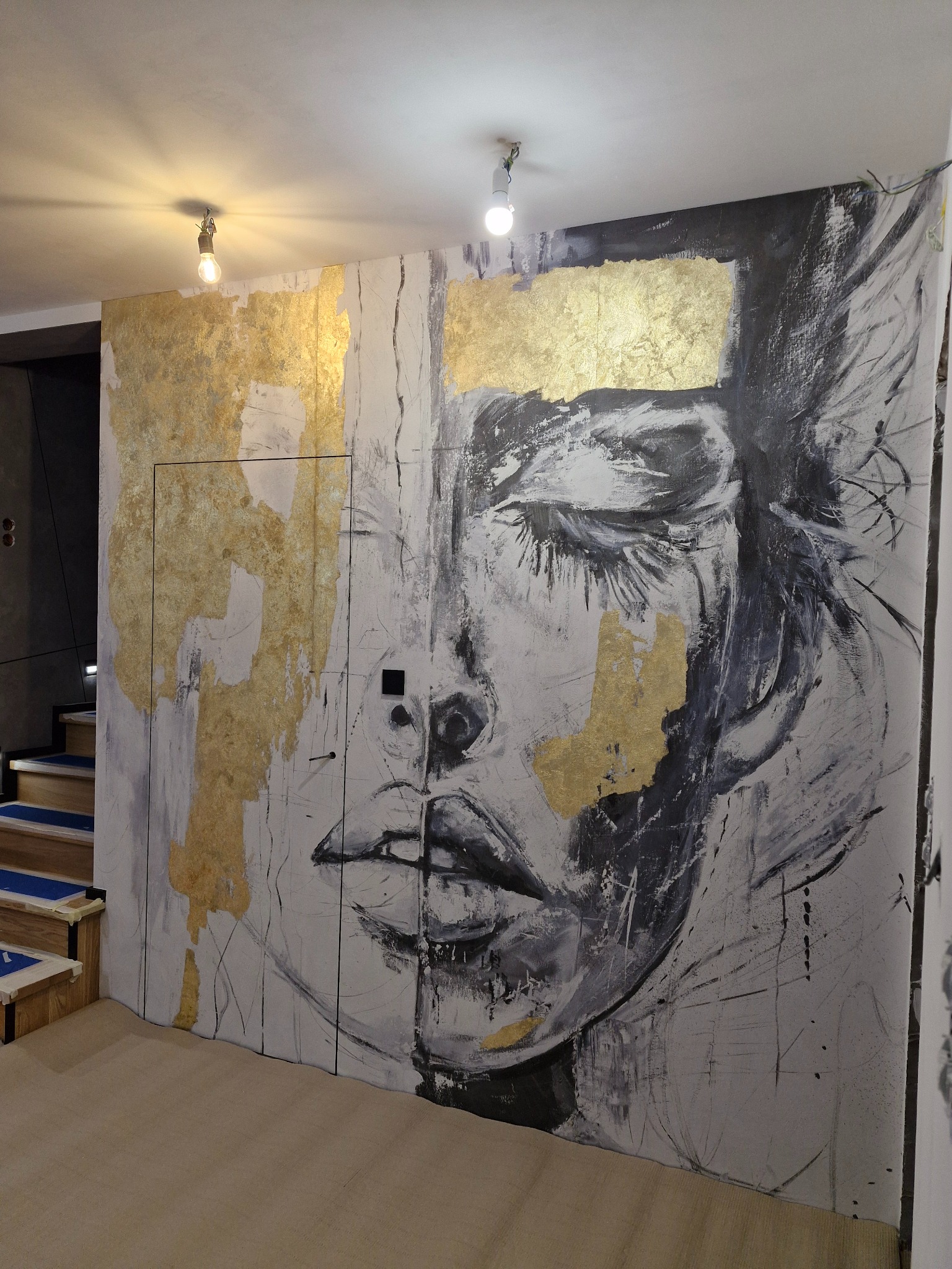 Abstrakcyjny mural z motywem twarzy kobiety, wykonany w odcieniach szarości i bieli z elementami złota, zdobiący ścianę z ukrytymi drzwiami w nowoczesnym wnętrzu.