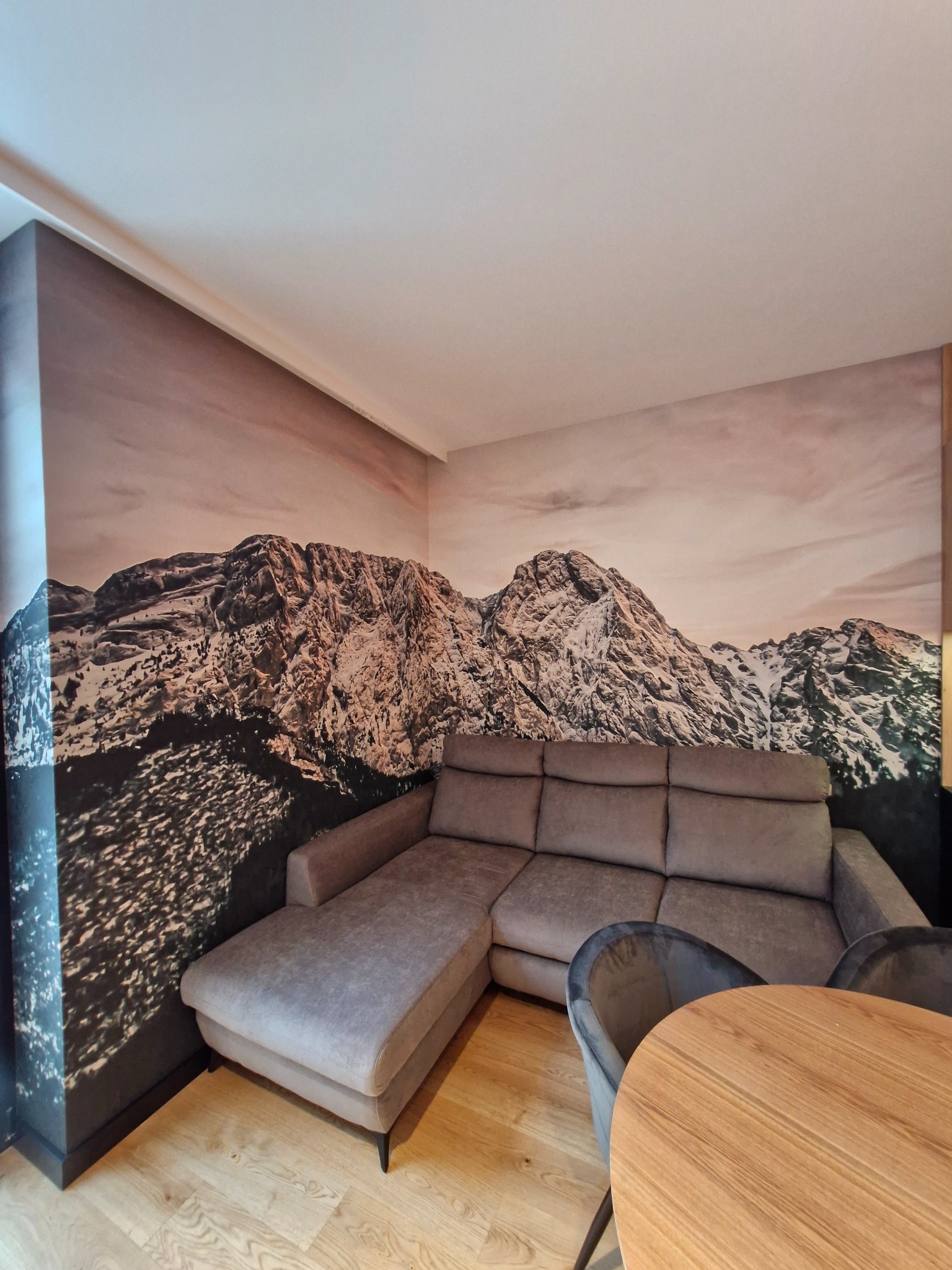 Salon z fototapetą górską, szary narożnik i drewniany stół. Fototapeta z motywem górskim na ścianie, tworząca spójną aranżację wnętrza.