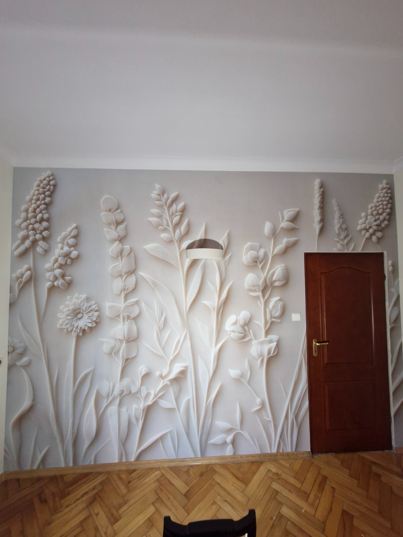 Ściana z dekoracyjną, trójwymiarową tapetą w kwiatowe wzory, obok ciemne drzwi. Parkiet na podłodze. Subtelne oświetlenie podkreśla detale.