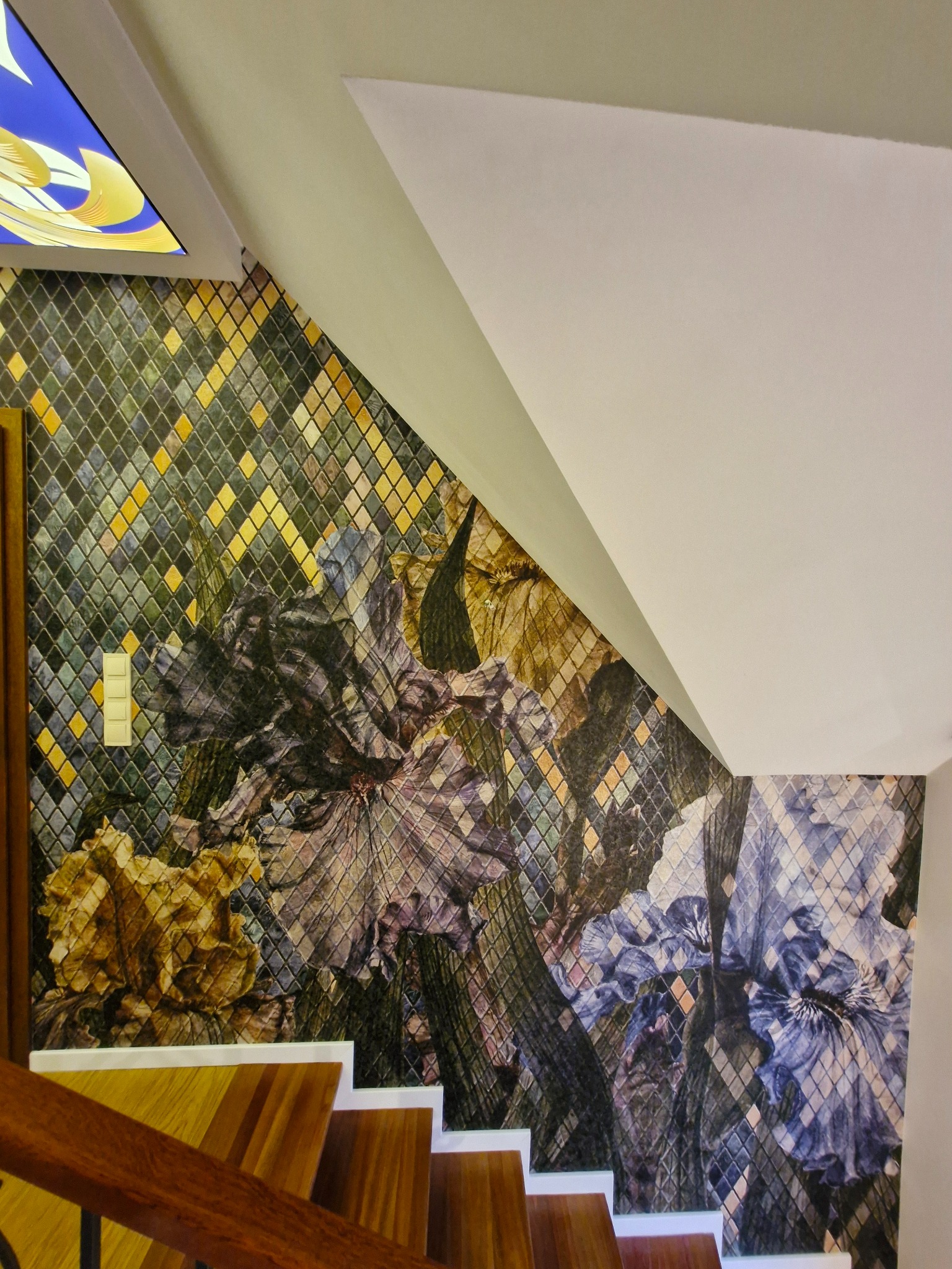 Ściana w klatce schodowej pokryta tapetą w mozaikę z motywem kwiatowym, widoczny fragment schodów z drewnianą barierką i włącznik światła.