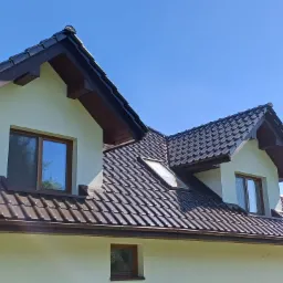 Fragment domu z jasną elewacją i brązowym dachem z dachówki, widoczne okna z brązowymi ramami i lukarna. Błękitne niebo w tle.