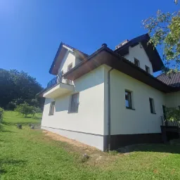 Jasny, otynkowany dom z ciemnobrązowym dachem i elementami wykończeniowymi, balkon z czarną balustradą, widok z dołu na tle zielonego trawnika i drzew, błękitne niebo.