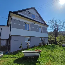 Dwukondygnacyjny dom z szarobiałą elewacją w poziome pasy, widziany z dołu, z wanną na trawniku i słonecznym niebem w tle.