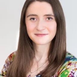 Portret uśmiechniętej kobiety z brązowymi włosami i fioletowym wisiorkiem na tle białej ściany.