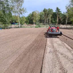 Czerwony traktor z glebogryzarką wyrównuje pole przygotowane pod trawnik na tle drzew.