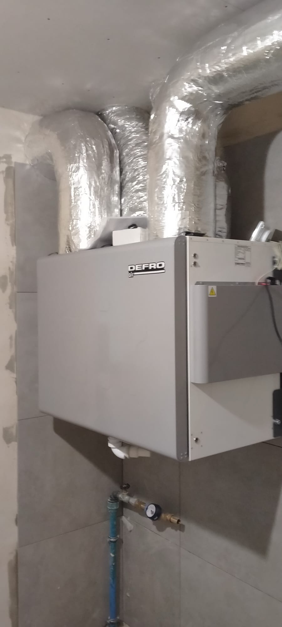 Szara jednostka centralna Defro Air z podłączonymi aluminiowymi przewodami wentylacyjnymi, zamontowana na ścianie z szarych płytek, widoczne również rury i manometr.