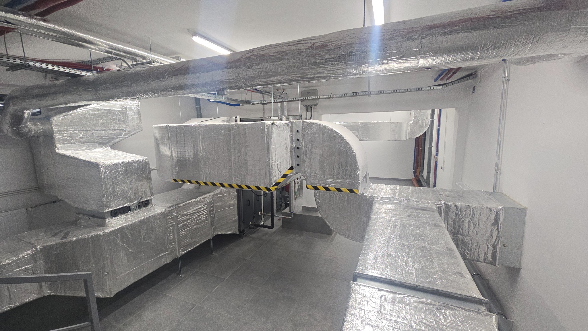 System kanałów wentylacyjnych owinięty folią izolacyjną w pomieszczeniu technicznym, z widocznymi czarno-żółtymi pasami ostrzegawczymi na niektórych elementach.