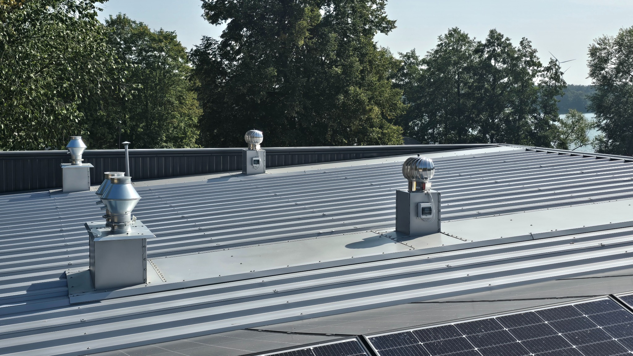 Szary blaszany dach z czterema kominkami wentylacyjnymi z turbiną wiatrową, panele słoneczne w prawym dolnym rogu, drzewa i jezioro w tle.