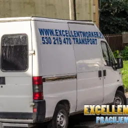 Biały bus transportowy firmy Excellent Worker z numerem telefonu i adresem strony internetowej, zaparkowany na tle zieleni.