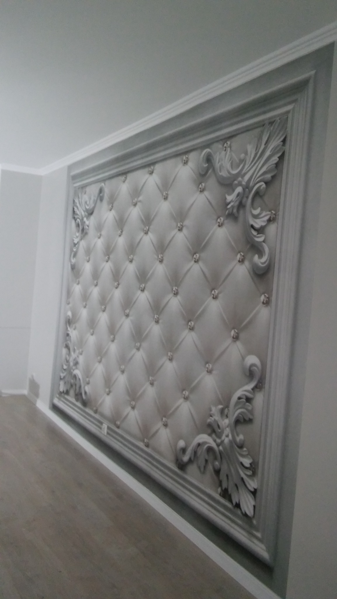 Ściana z dekoracyjnym panelem tapicerowanym w ramie z ornamentami i kryształkami, widok pod kątem.