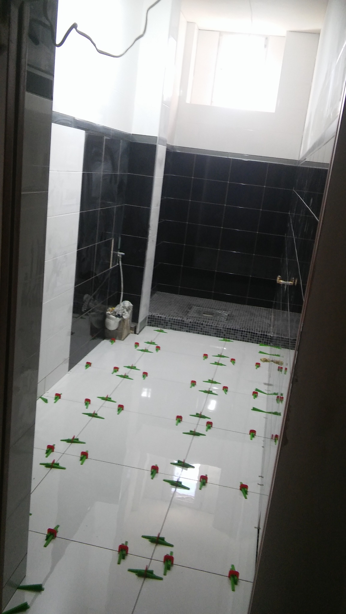 Białe płytki podłogowe w trakcie układania w łazience z czarnymi płytkami ściennymi, widoczne krzyżyki dystansowe i wiadro z narzędziami w rogu.