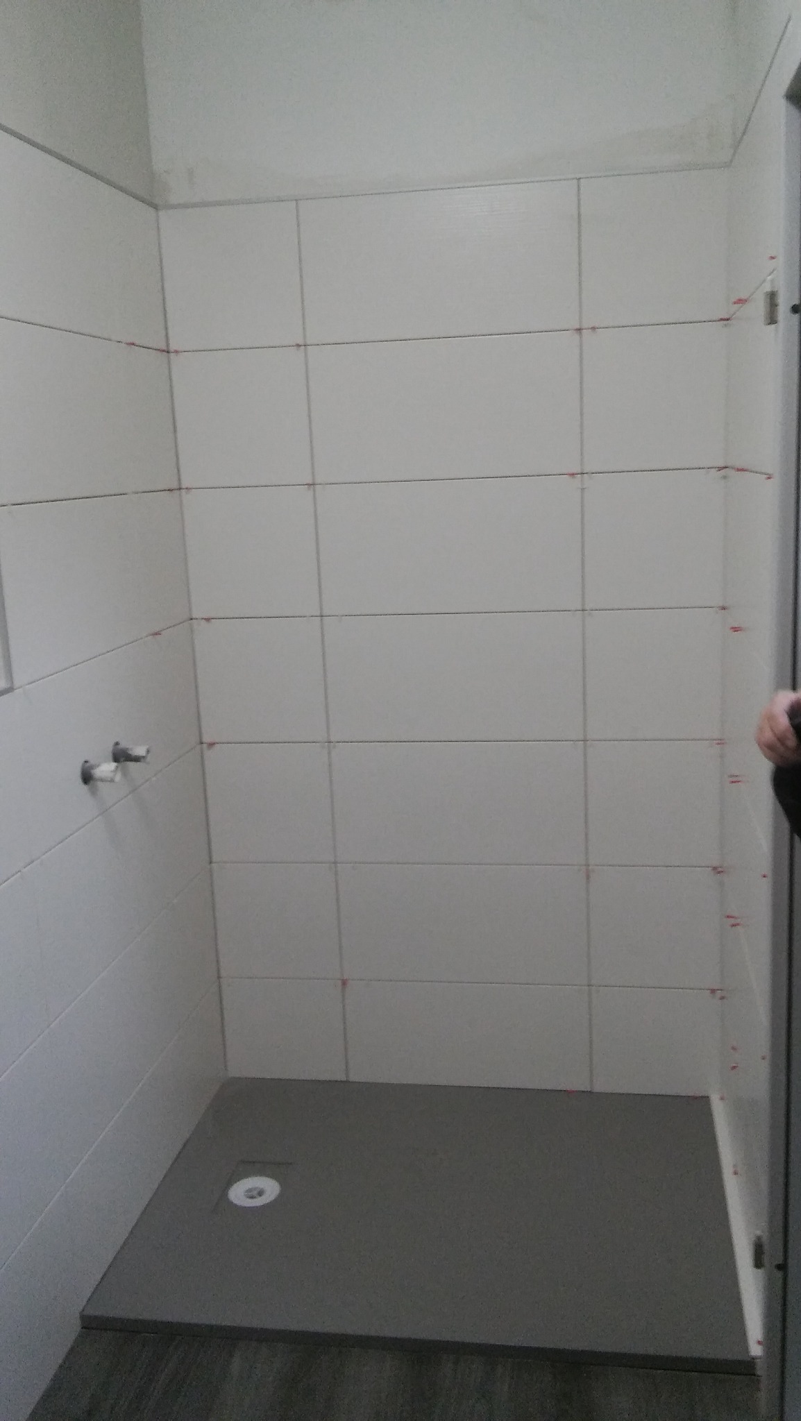 Świeżo wykafelkowana kabina prysznicowa z białymi płytkami, widoczne czerwone krzyżyki dystansowe, szary brodzik z odpływem, ujęcie z góry w trakcie prac wykończeniowych.