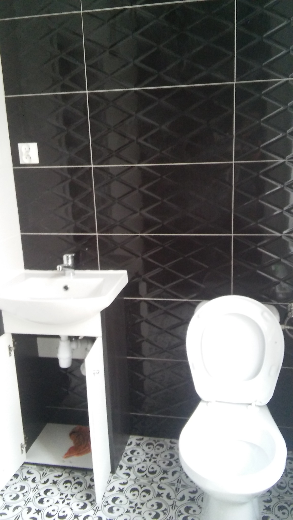 Nowoczesna łazienka z czarnymi, wzorzystymi płytkami na ścianach, białą umywalką z szafką i otwartymi drzwiczkami oraz toaletą na podłodze w biało-czarne wzory.
