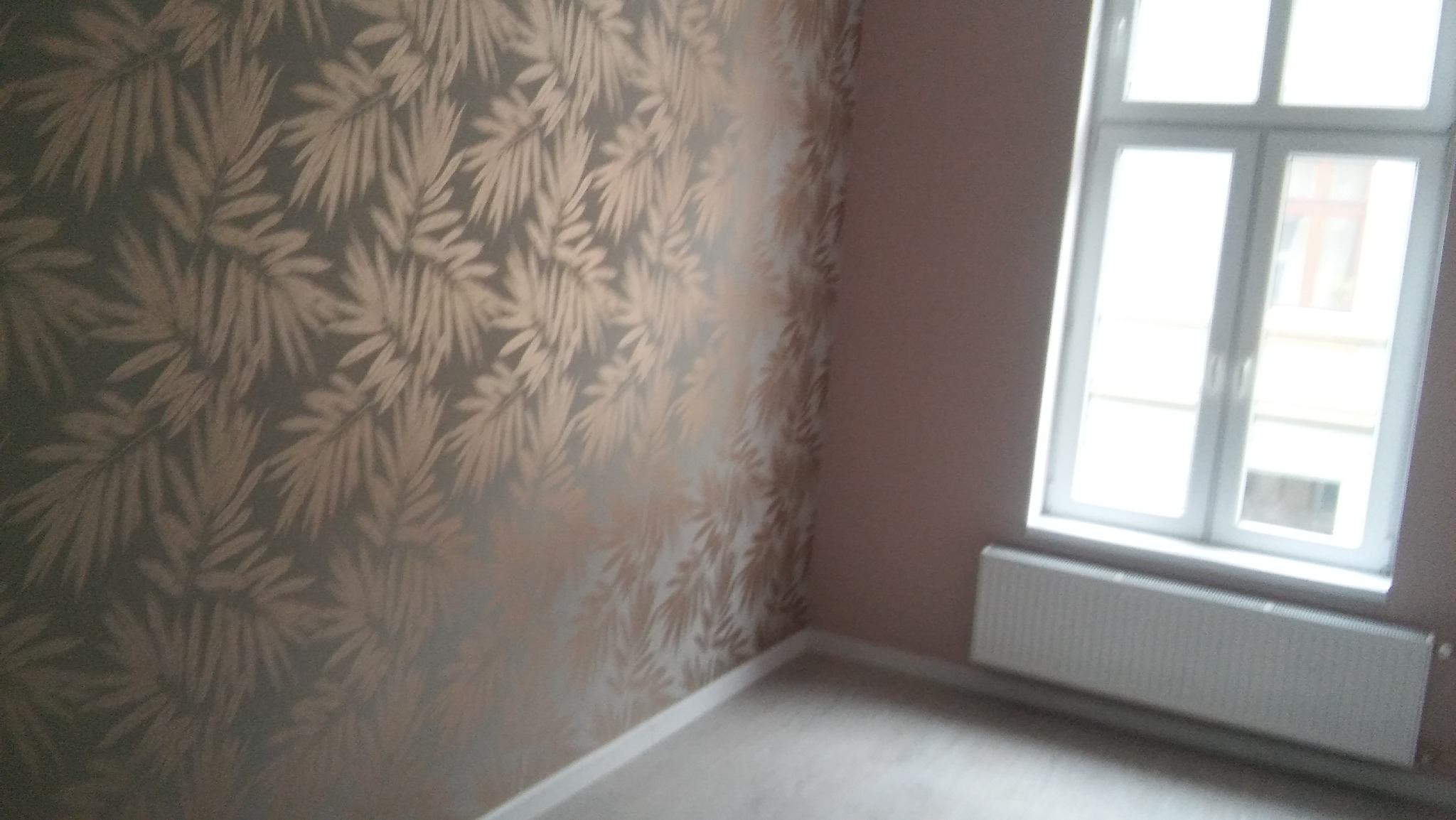 Pokój po remoncie: ściana pokryta tapetą w złote liście na szarym tle, białe okno z grzejnikiem pod spodem, jasna podłoga.