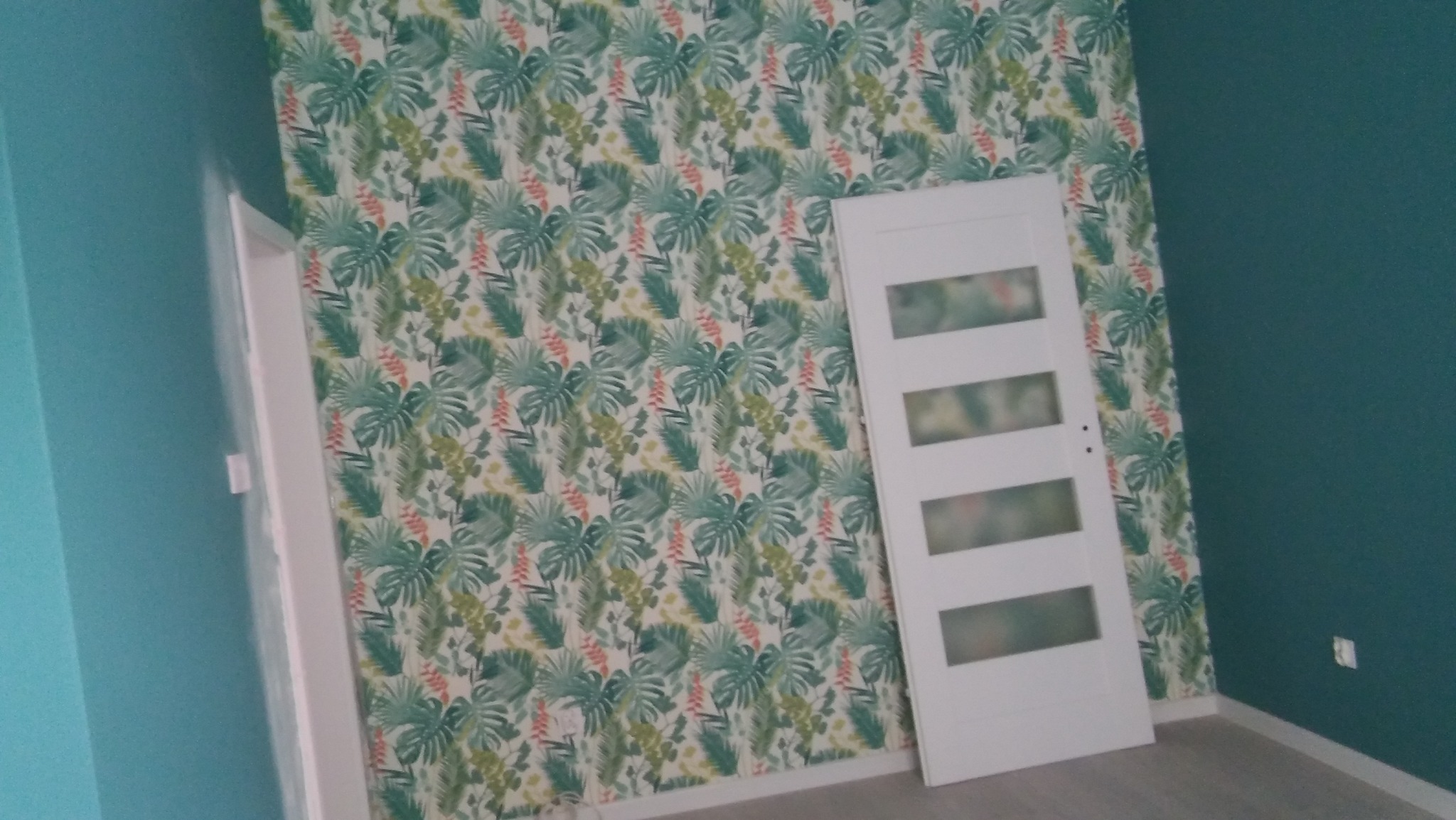 Pokój po odświeżeniu z turkusowymi ścianami i tapetą w tropikalne liście, białe drzwi z przeszkleniami opierają się o ścianę.