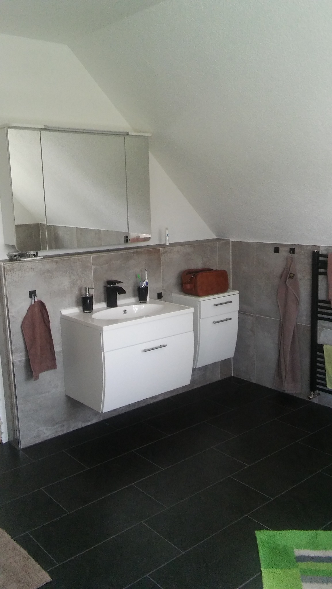 Nowoczesna łazienka na poddaszu z czarną armaturą, białą szafką pod umywalką z szufladami i lustrem w szafce, szare płytki na ścianach i czarna podłoga z płytek.