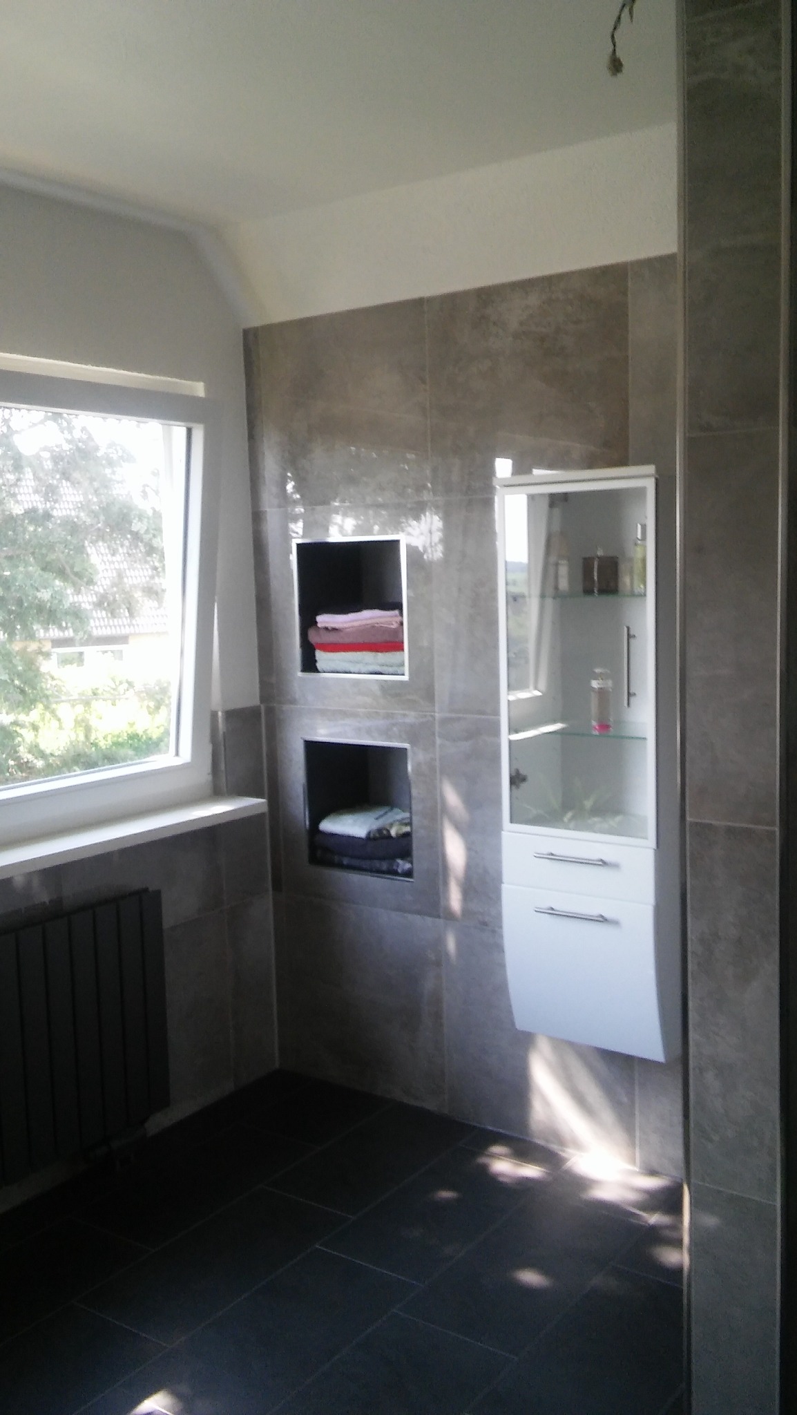Wykończona łazienka z szarymi płytkami, wbudowanymi półkami z ręcznikami i białą szafką z lustrzanymi drzwiczkami, czarne płytki podłogowe, grzejnik pod oknem.