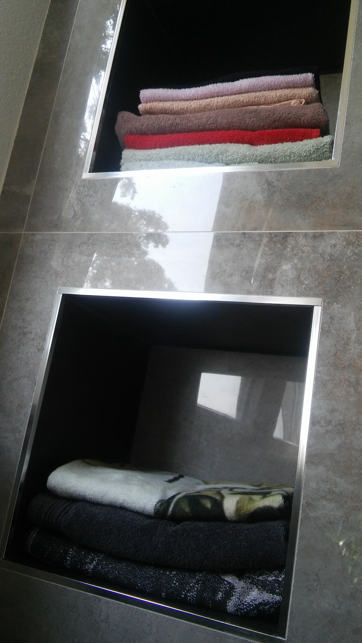 Dwa wnękowe schowki w łazience wyłożonej szarymi płytkami, z widocznymi stertami ręczników w różnych kolorach i wzorach.