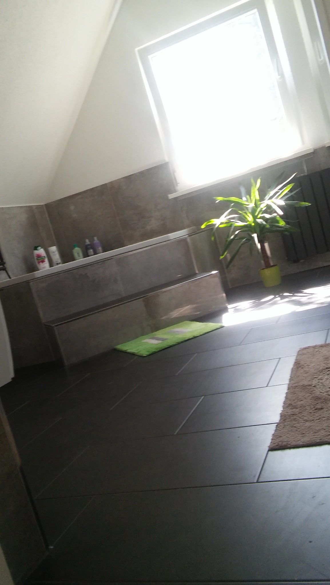 Jasna łazienka na poddaszu z ciemnymi płytkami podłogowymi, dwupoziomową zabudową wanny wykończoną płytkami w odcieniach szarości, zielonym dywanikiem i kwiatem doniczkowym w żółtej osłonce...