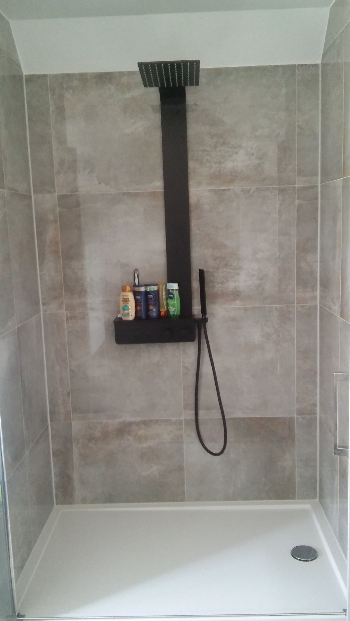 Nowoczesny prysznic z czarną deszczownicą i półką na kosmetyki, wykończony beżowymi płytkami.