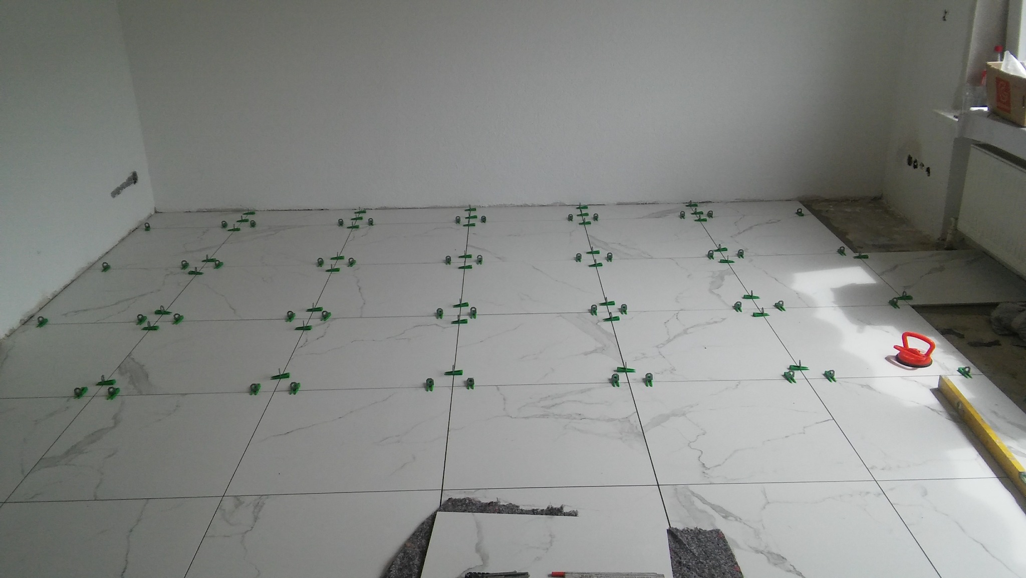 Białe płytki podłogowe z szarym wzorem żyłkowania, ułożone w rzędach z użyciem zielonych klipsów poziomujących, widoczny fragment niezakończonej podłogi przy ścianie, poziomica i przyssawka...