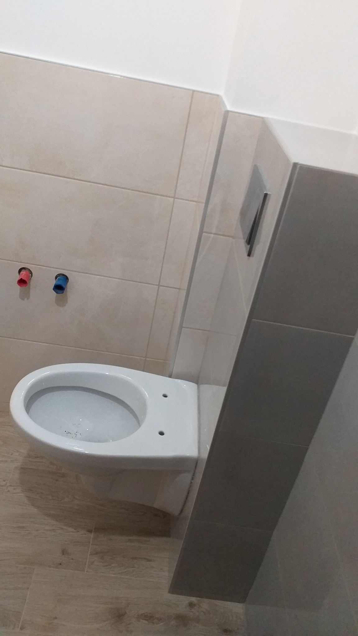 Biała, wisząca toaleta zamontowana w łazience z beżowymi płytkami, widoczne rury doprowadzające wodę z niebieskim i czerwonym oznaczeniem.