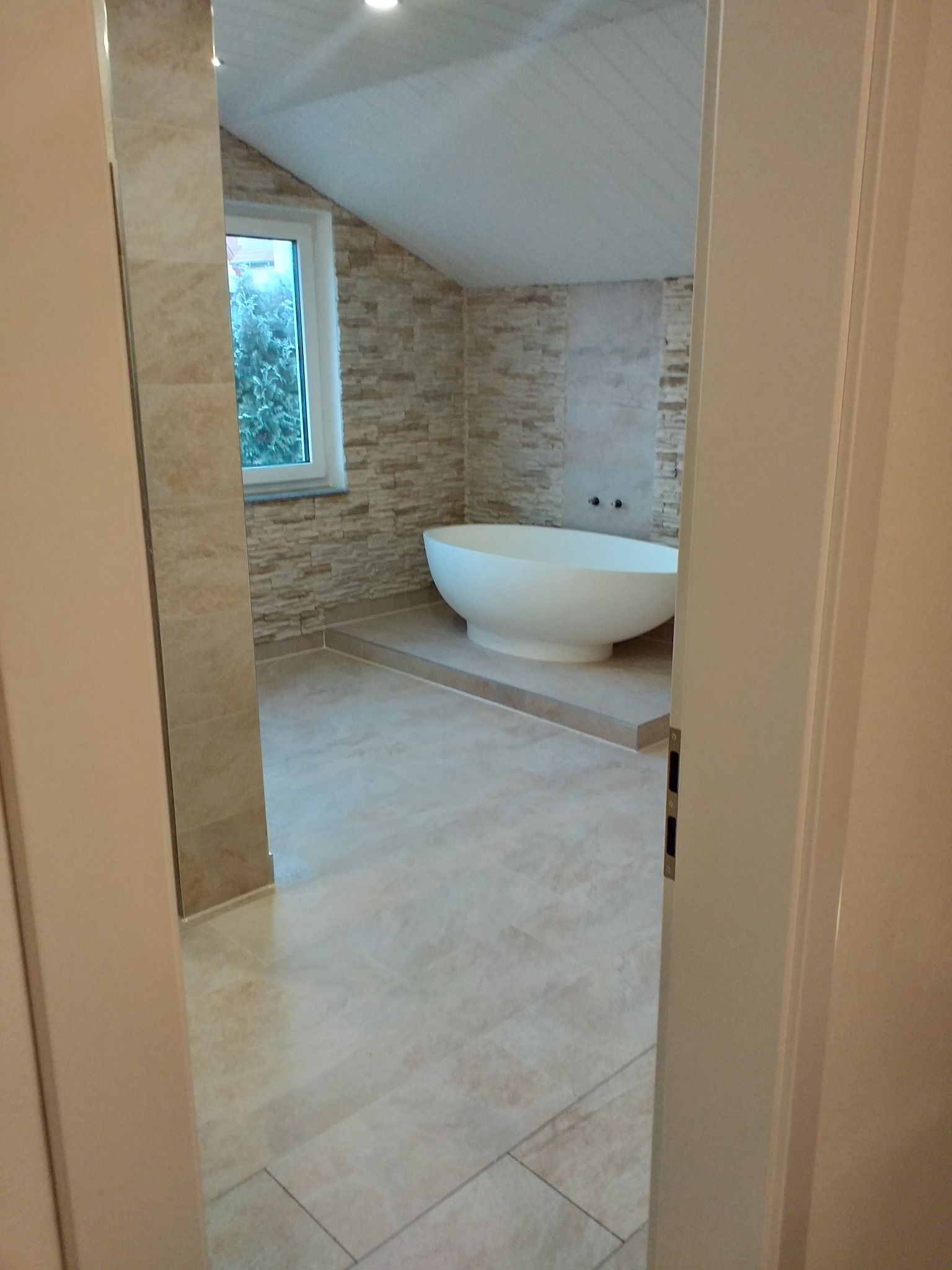 Nowoczesna łazienka z wolnostojącą wanną, wykończona płytkami w odcieniach beżu i szarości, widok przez uchylone drzwi.