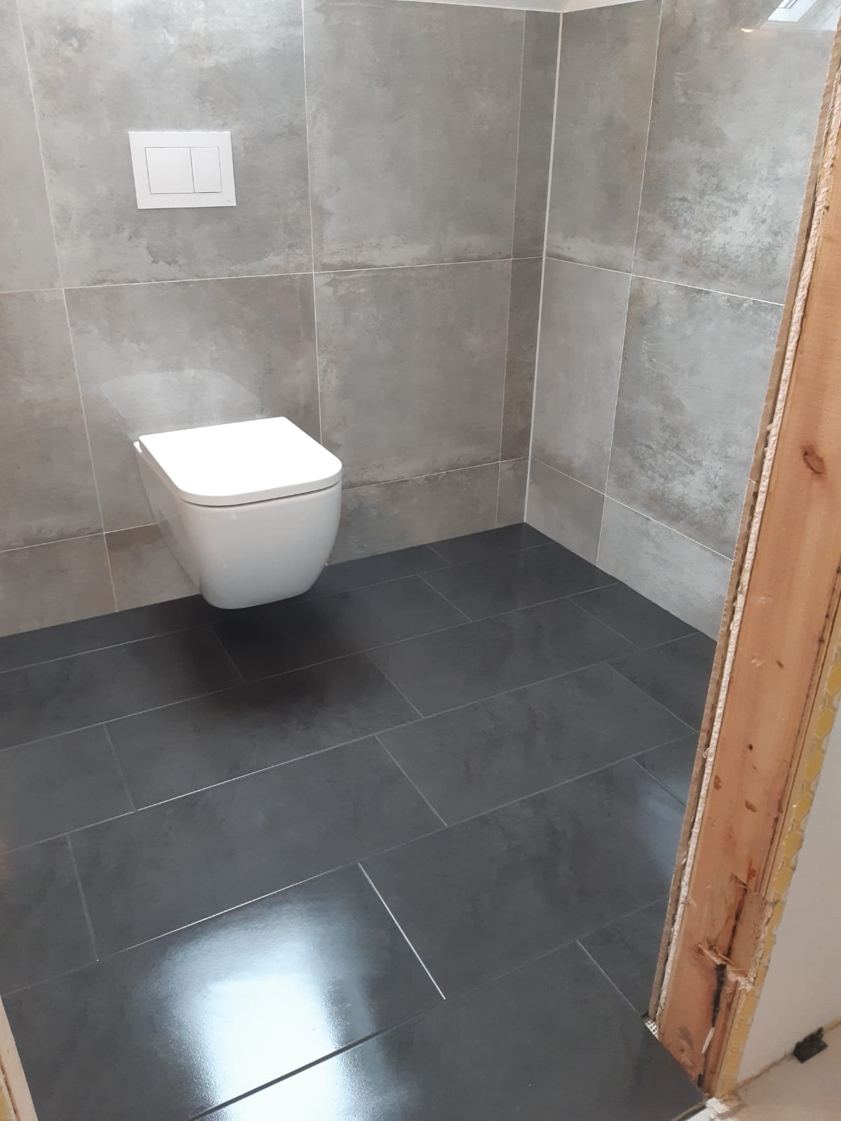 Nowoczesna łazienka z podwieszaną toaletą, wykończona szarymi płytkami na ścianach i podłodze, z widoczną surową ościeżnicą drzwi.