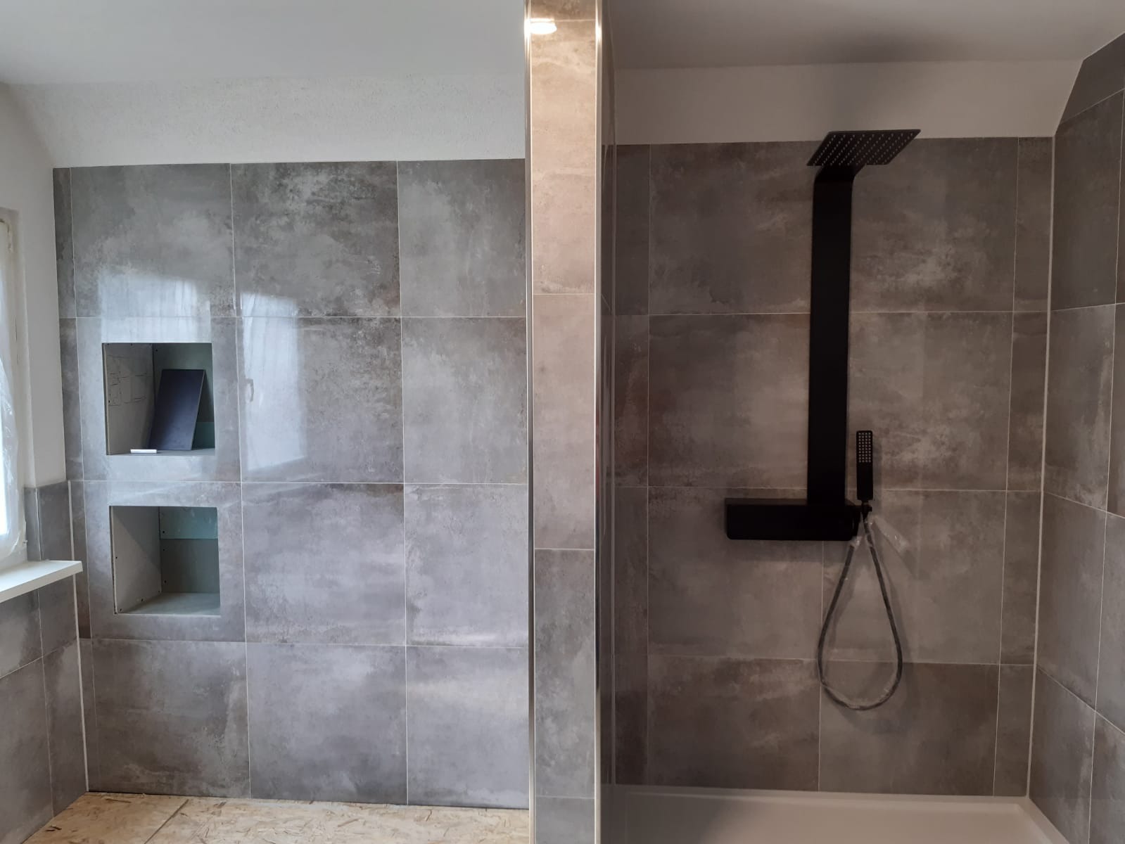 Nowoczesna kabina prysznicowa z szarymi płytkami imitującymi beton, czarną deszczownicą i wnękami w ścianie.