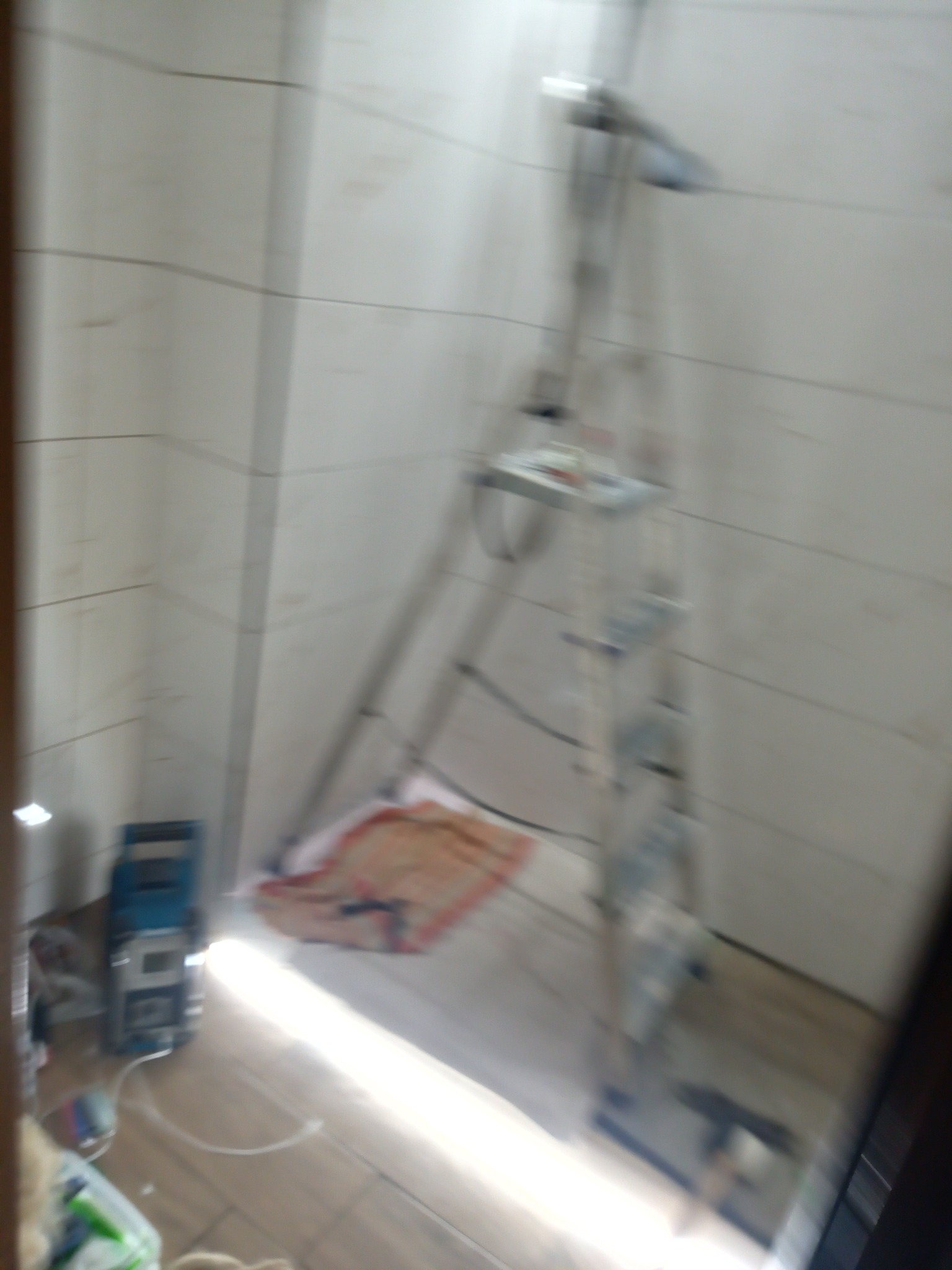 Rozmazane ujęcie wnętrza w trakcie prac: drabina aluminiowa oparta o ścianę z białych płytek, niebieska skrzynka narzędziowa na podłodze. Widoczny fragment podłogi i ręcznik.