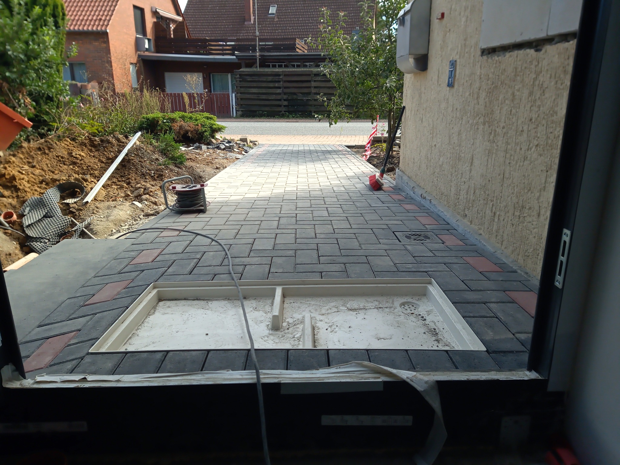 Ułożona kostka brukowa w odcieniach szarości i czerwieni prowadząca do budynku, widoczny fragment studzienki kanalizacyjnej z osadnikiem, kabel elektryczny zwinięty na bębnie leży obok wykopu...